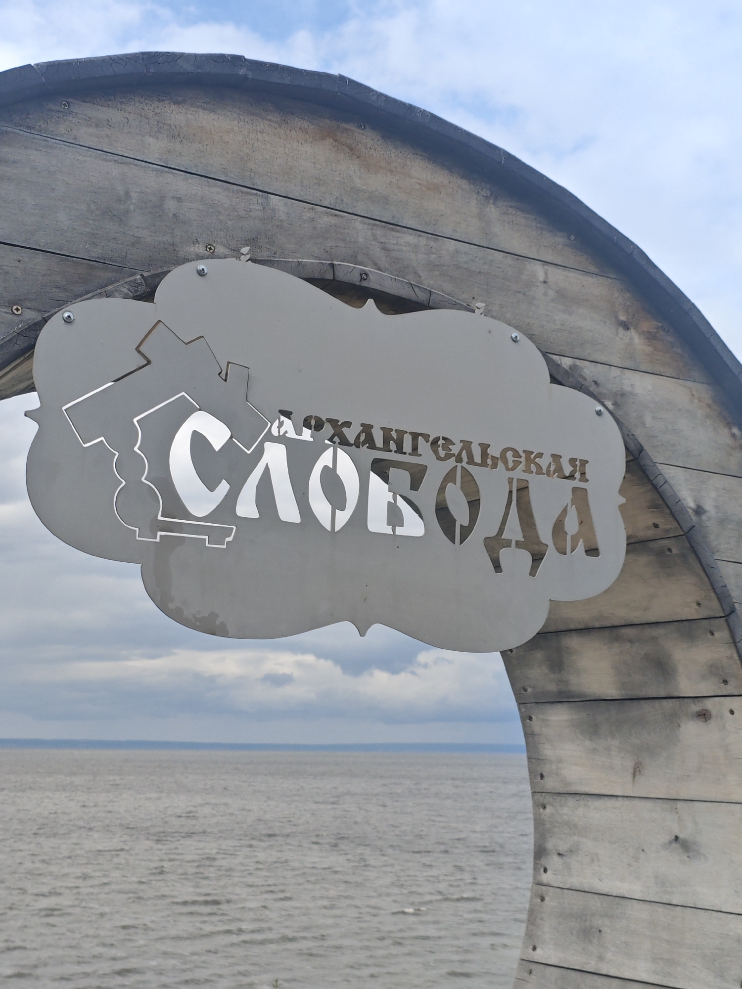 An outdoor metal sign with cut-out text is attached to a curved wooden structure. The sign is shaped like a stylized cloud with a gear graphic on the left side. The text on the sign reads "АРХАНГЕЛЬСКАЯ СЛОБОДА". The sign is set against a background of the sky and a large body of water. The wooden structure appears to be part of an archway or entrance. The scene is likely located in Arkhangelsk, Russia, as indicated by the text on the sign.