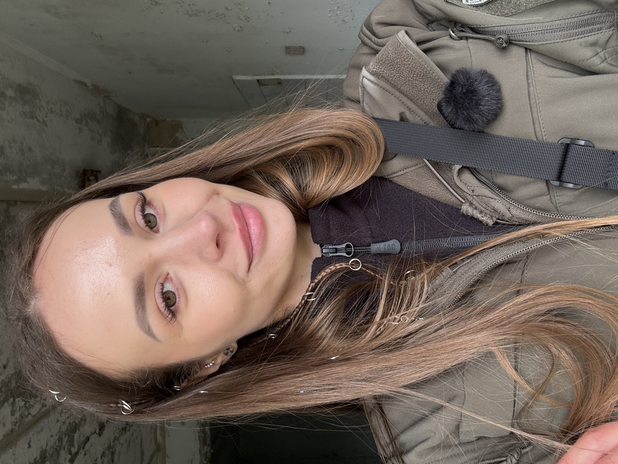 A fair-skinned woman with light brown hair stands in an unidentified location in the city of Prypiat, Ukraine. The location appears to be indoors in a building, possibly abandoned, with concrete walls and poor lighting visible behind her.
She wears a khaki-colored jacket. A black strap of material, possibly a bag strap, runs diagonally across her body. A small black fluffy object is attached near her shoulder on the jacket. She is close to the camera. Her hair is long with silver rings adorning her hair and small braids. She is wearing earrings. She is looking at the camera and has a slight smile.