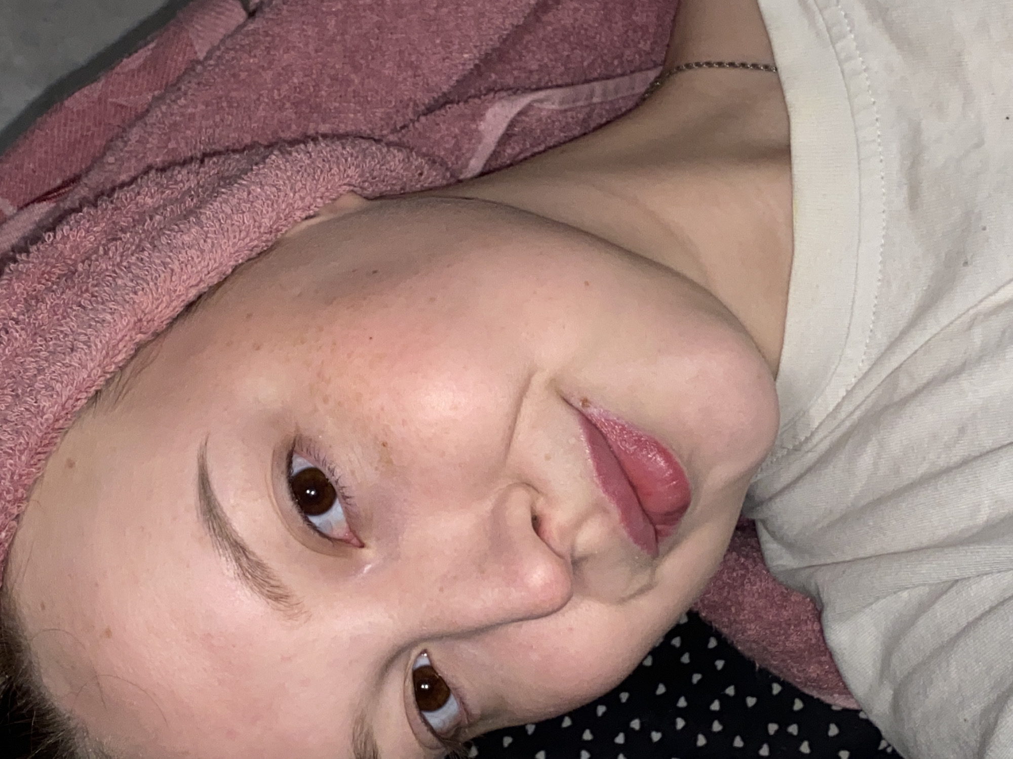A person lies with a pink towel wrapped around their head, wearing a white shirt and a silver necklace. The background shows a black surface with white polka dots. The person has brown eyes and freckles. The image does not provide information about the location or the city of Chaikovsky, Russia.