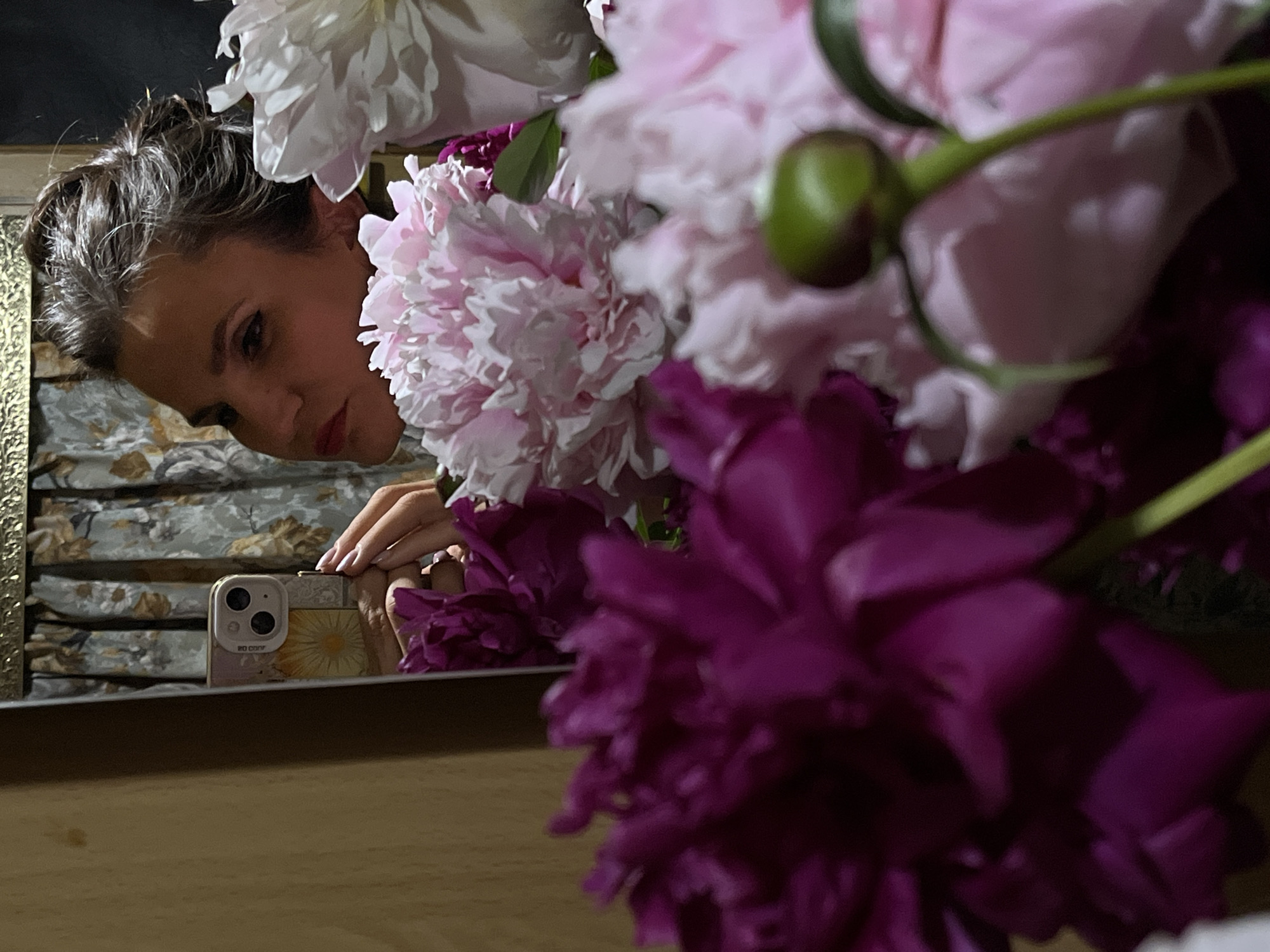 This is a medium shot, eye-level, indoor, selfie-style photograph taken in Petropavlovsk-Kamchatsky, Russia. The photo captures a woman taking a selfie in a mirror. In the foreground, a bouquet of peonies is arranged, with vibrant purple and soft pink flowers dominating the frame. A small green bud is visible in the lower right corner. Reflected in the mirror, the woman's face is angled to the left, with her dark hair pulled back. She is holding a smartphone, which is visible in the mirror, and her manicured hand is reaching out. Behind her, a floral curtain is visible, and to the left, a portion of a wooden surface can be seen. The overall lighting suggests an indoor environment, possibly a home.