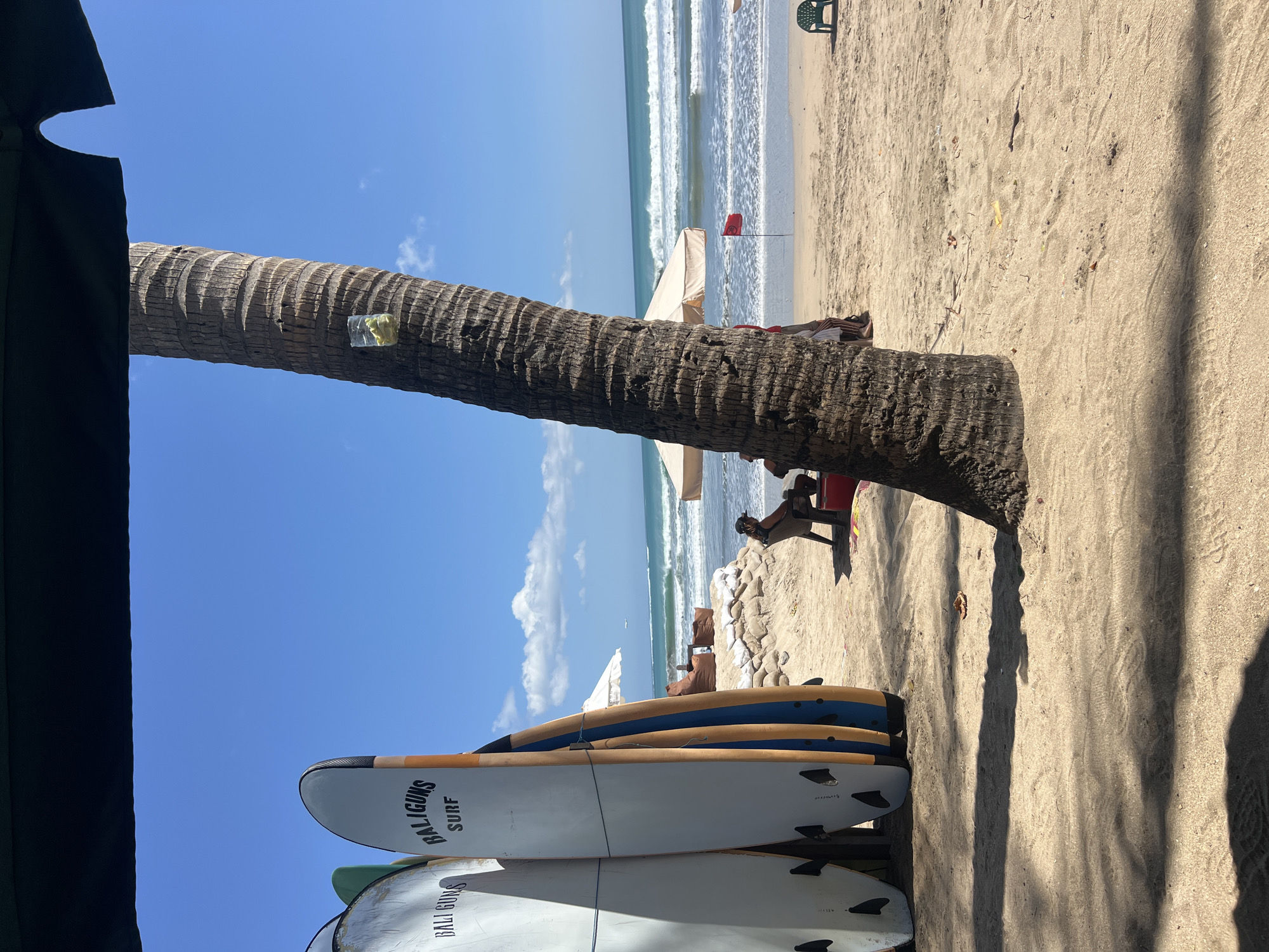 The image depicts a sunny day at Kuta Beach, Indonesia, with a focus on the sandy shore and the ocean. In the foreground, a thick palm tree trunk dominates the right side of the frame, with a clear plastic cup containing light-colored items, possibly fruit, attached to it. To the left, three surfboards are propped up, two with "BALI GUNS SURF" visible on them. Beyond the surfboards, the beach stretches towards the water, where gentle waves are rolling in.
Several people are present on the beach. A person is sitting in a chair facing the ocean, appearing relaxed. In the middle ground, a white umbrella provides shade. A red flag is planted in the sand near the water's edge, indicating a possible warning or marker. Further back, more umbrellas and beach chairs are scattered. The sky is a vibrant blue with scattered white clouds, and a plane is visible in the distance. The overall scene suggests a typical beach day with recreational activities.