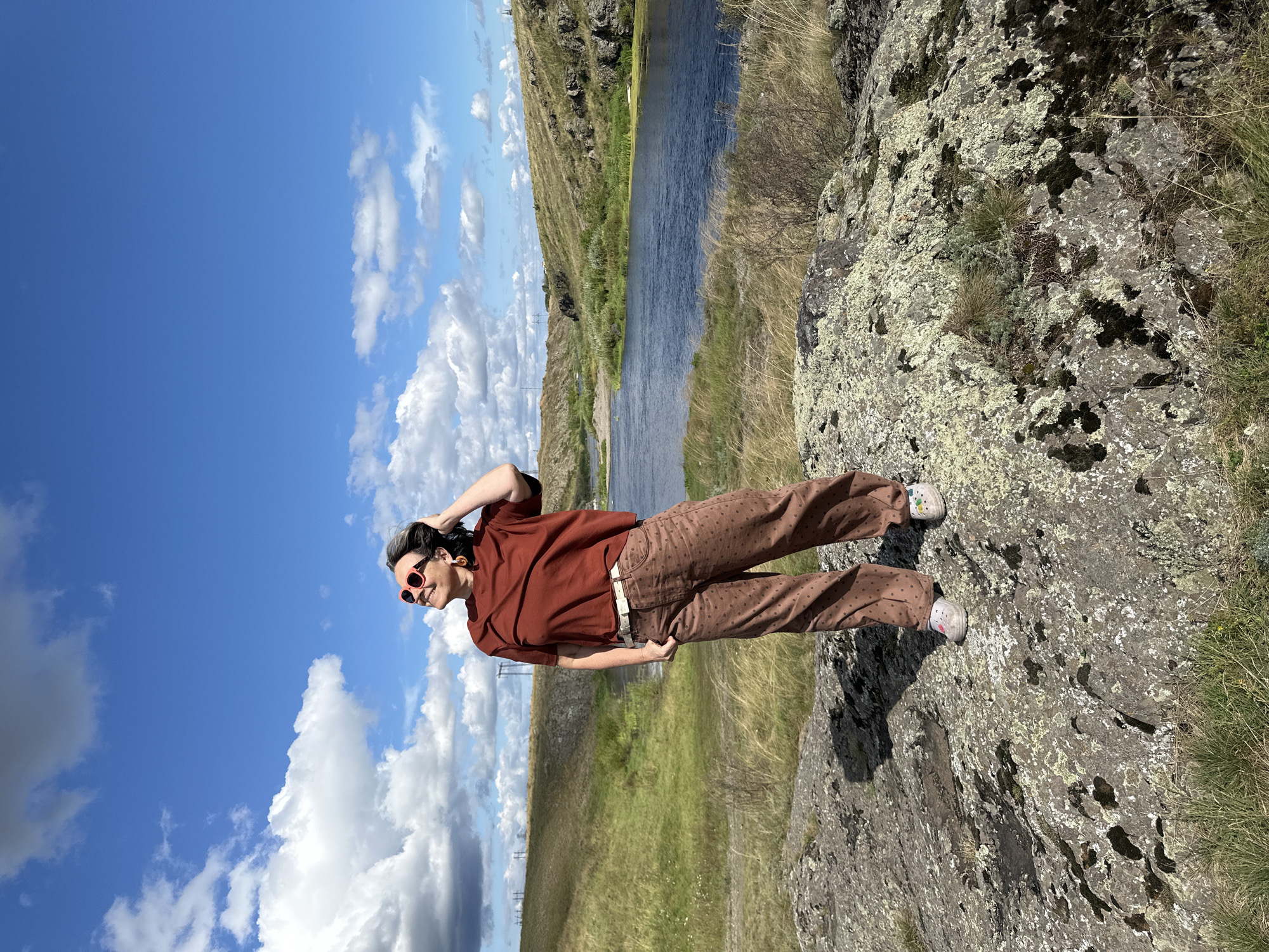 A medium shot captures a woman standing on a rocky outcrop. She is wearing a reddish-brown short-sleeved shirt, brown polka-dot pants, a white belt, and white sneakers. She has dark hair, wears red-framed sunglasses, and large orange earrings. The woman is looking up and to her left, with her right hand touching her hair.
The background features a blue sky with scattered white clouds, a body of water, and rolling grassy hills. The landscape appears natural and undeveloped, with some rocky formations and vegetation. In the distance, power lines and poles are visible on the horizon. The lighting suggests it is a sunny day.