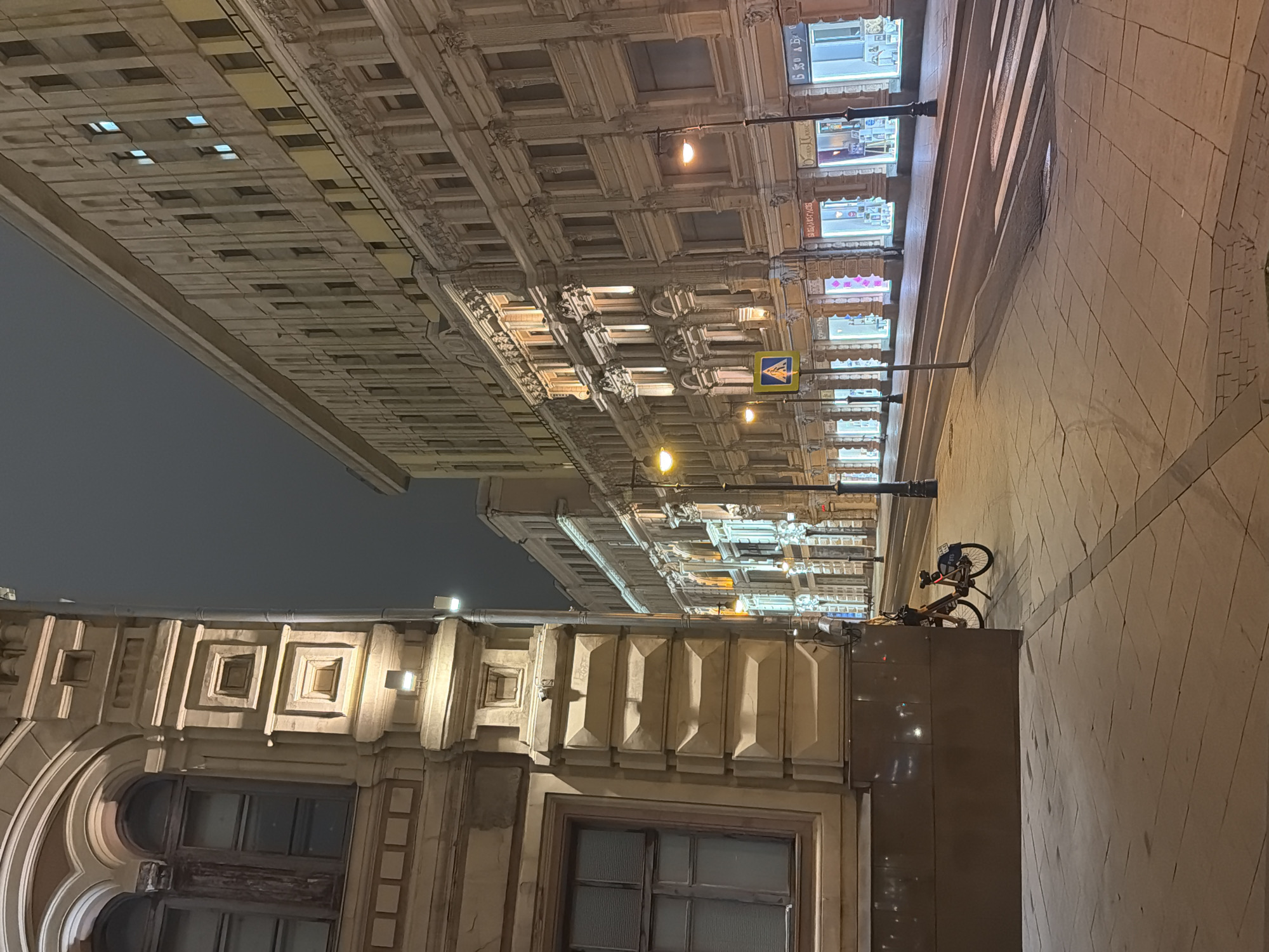 The image depicts a street scene at night in Moscow, Russia. Two buildings frame the view. The building on the left has a light-colored façade with detailed architectural elements, including windows with framed panes and a series of squared and rectangular shapes. The building on the right has a more ornate façade, featuring intricate patterns and illuminated windows. Street lamps are visible along the sidewalk, with a pedestrian crossing sign in the middle. A bicycle is parked alongside the building on the right. The sky is dark. The street is paved with light-colored stone blocks. The overall ambiance is illuminated and appears well-maintained.