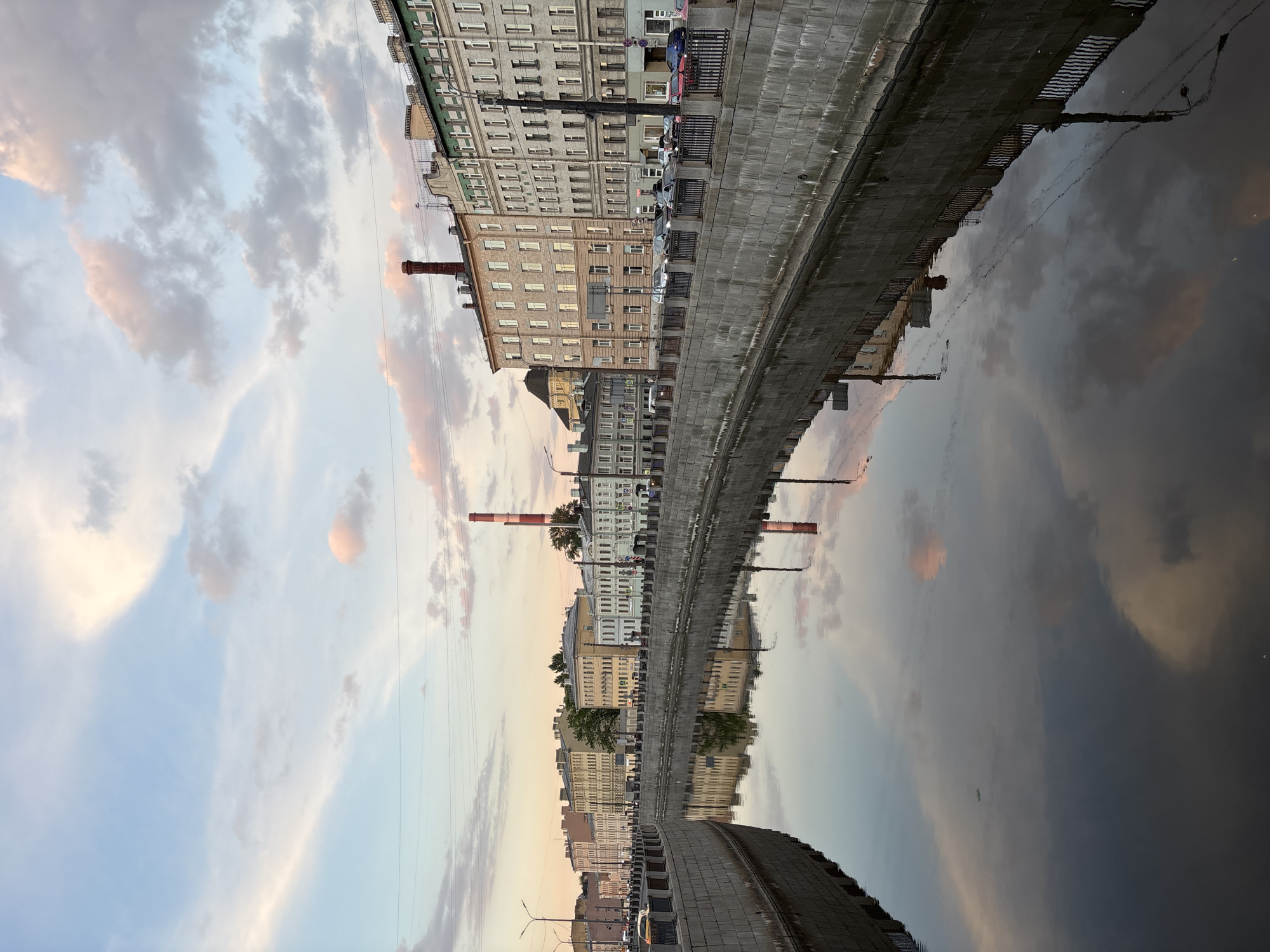 The image captures a serene urban canal scene, likely at dawn or dusk, characterized by the soft, diffused light. The sky, occupying the upper portion, displays a mix of light blue and grey clouds streaked with warm pink and orange hues, indicative of either sunrise or sunset.
Along the left bank of the canal, a continuous stretch of multi-story buildings stands, featuring varied architectural styles with numerous windows and facades in tones of grey, beige, and light green. A solid stone or concrete embankment separates the buildings and a parallel street from the water. Several cars are parked along this street. Prominently rising above the buildings are two tall industrial chimneys; one is a distinctive red and white striped smokestack, and another appears to be made of brick. Utility wires are faintly visible against the sky.
The calm, dark water of the canal beautifully reflects the sky, clouds, and the buildings lining its banks, creating an almost symmetrical, mirror-like effect for the lower half of the image. The reflections include hints of trees and streetlights. No discernible people or text are visible in the photograph, contributing to a quiet, contemplative atmosphere. The scene evokes a sense of peace in an urban environment.