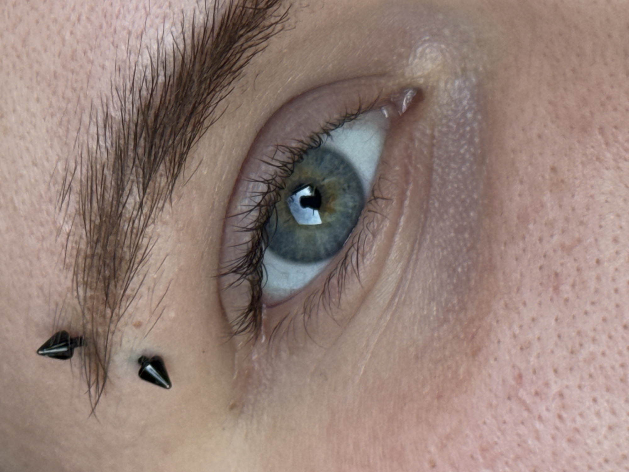 This is a close-up, horizontal, outdoor, natural light photograph of a person's left eye and eyebrow. The subject's skin appears fair and has visible pores. The eyebrow is dark and bushy. Two black, spike-shaped piercings are visible in the eyebrow area. The eye itself is a striking blue-green color with dark eyelashes. The iris shows a reflection of a light source. The overall tone of the image is neutral, focusing on the details of the face and piercings. The setting is implied to be an everyday environment given the natural lighting and lack of specific background elements. There is no visible text in the image.