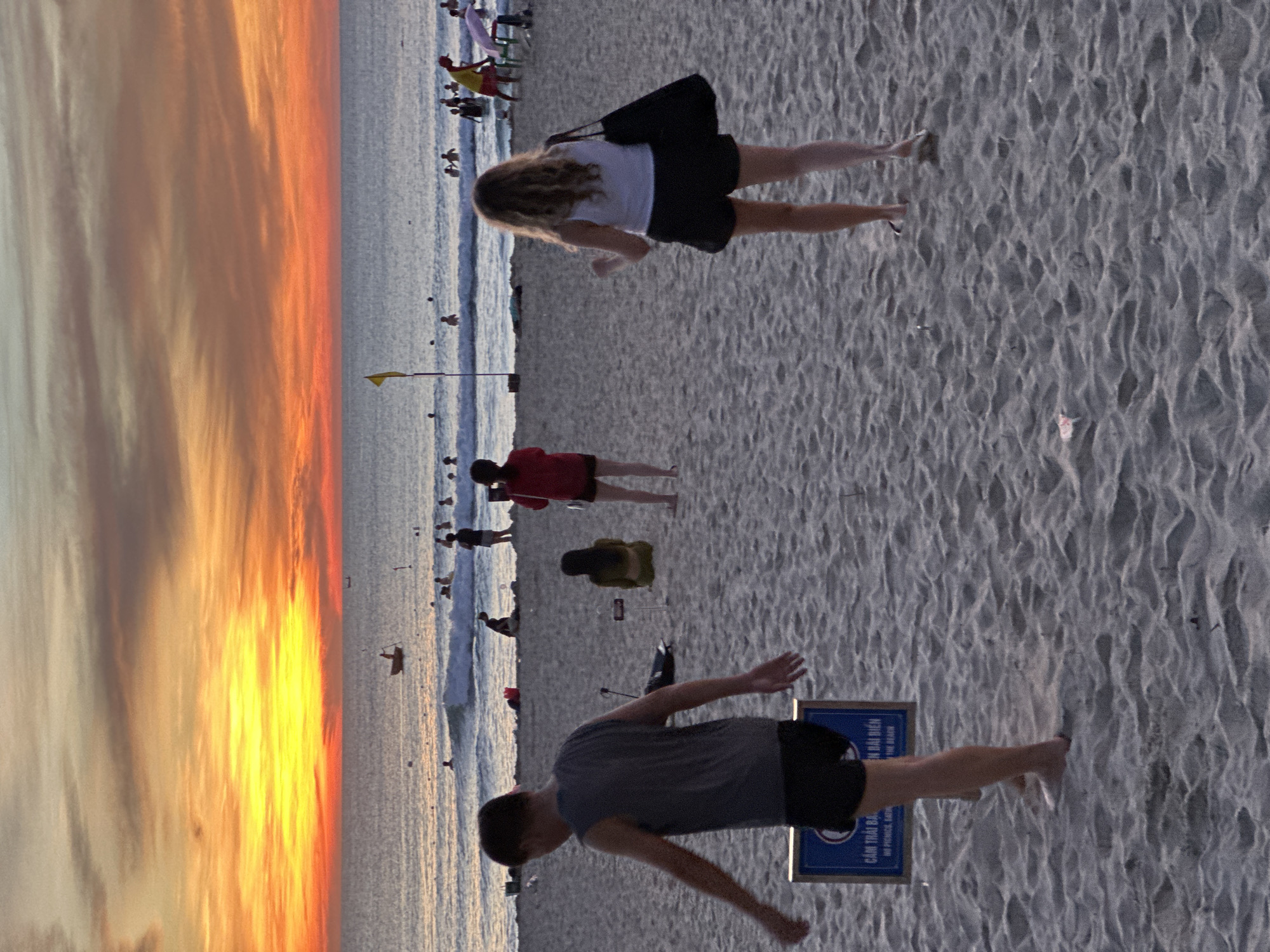 The image depicts a lively beach scene at sunset in Đà Nẵng, Vietnam. The sky is ablaze with vibrant hues of orange, yellow, and pink, reflecting on the calm ocean. In the foreground, two people are walking on the sandy beach. A man in a grey tank top and black shorts is walking away from the camera, while a woman in a white top and black shorts is walking towards the camera. Both appear to be enjoying a leisurely stroll.
In the background, several other people are seen wading in the shallow waters or relaxing on the beach. A small boat is visible in the distance, adding to the scenic view. The overall atmosphere is one of relaxation and enjoyment, with the warm sunset casting a peaceful glow over the beach. The presence of a lifeguard and beach chairs suggests that this is a popular spot for tourists and locals alike. The sign in the foreground, with Vietnamese writing, indicates a public beach in Đà Nẵng.