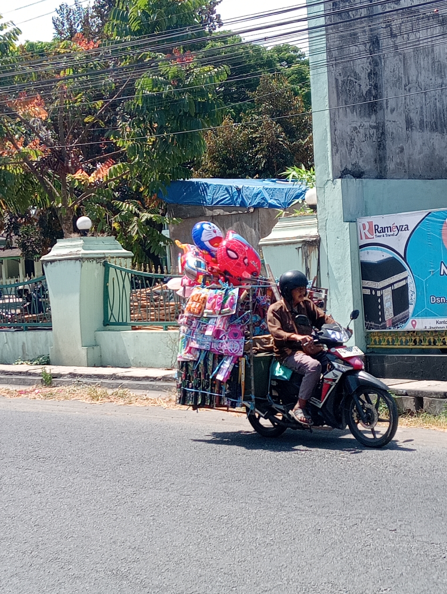 A man wearing a brown long-sleeved shirt, dark pants, and a black helmet rides a motorcycle laden with balloons and merchandise. The motorcycle is black with red and white accents. Attached to the rear of the motorcycle is a large rack displaying numerous colorful balloons, including some shaped like cartoon characters and superheroes, along with packages of small items, possibly toys or accessories. The man is wearing sandals. He is riding on a paved road, likely in an urban or semi-urban area.
In the background, there is a light green fence and a light blue tarp covering something. A building with a textured, weathered gray wall and light green trim is visible to the right. A sign on the building advertises "Raméyza Tour & Travel" with an image of the Kaaba. Trees with green foliage and some reddish-orange flowers are also present in the background. Power lines are strung across the scene. The ground is asphalt, with a dry, grassy verge along the roadside. The lighting suggests it is daytime, with bright sunlight casting shadows. This scene is consistent with street vending or mobile sales in a location such as Jabon, Indonesia.