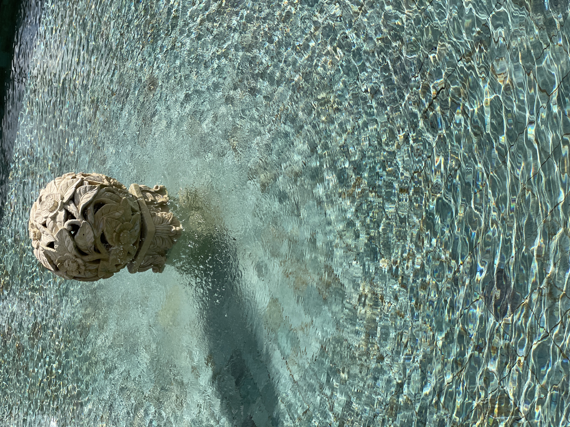 A high-angle, natural light shot shows a stone ball fountain in a pool. The pool's surface is textured with ripples. The stone ball, positioned on the left, is carved with a floral design. Its shadow is visible on the pool floor. The location of the scene is not discernible.