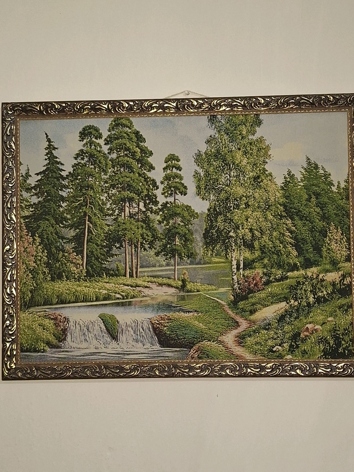The image shows a framed tapestry depicting a serene natural landscape. The scene features a gentle waterfall flowing into a calm body of water, surrounded by lush green foliage and tall trees. A winding dirt path leads through the grassy terrain, suggesting a park or natural reserve. The sky is a pale blue with some cloud cover. The tapestry is ornate and appears to be a decorative piece. There are no people visible in the tapestry itself. The tapestry is hung on a plain white wall. The image does not contain any recognizable landmarks or specific features that would allow for identification of its location within Almaty, Kazakhstan.