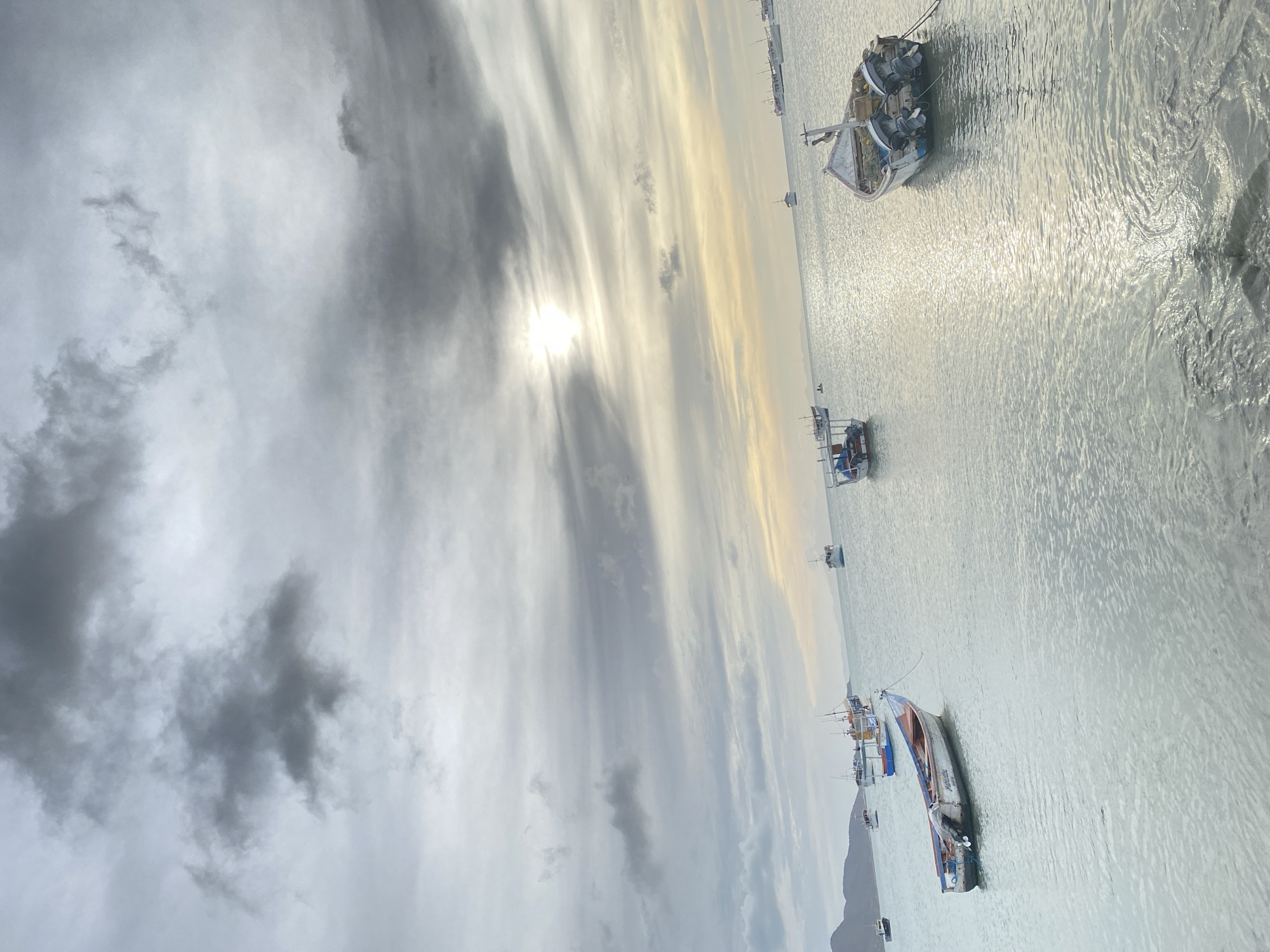 This image depicts a marine scene in Juan Griego, Venezuela, under an overcast sky with visible sunlight. The foreground shows calm water with a strong sun glare reflecting on its surface, indicating the sun's position. Approximately twenty-five small to medium-sized fishing vessels are distributed across the water.
In the immediate right foreground, a white and blue fishing boat with two outboard motors is moored, exhibiting visible weathering and various items stored within its hull. To the left, a white boat with red and blue trim, named "NORIEGA" on its side, is also moored. Further back, numerous other boats, primarily white, are scattered across the water. Many possess small cabins or canopies.
The sky, occupying the upper two-thirds of the frame, is heavily covered with stratocumulus clouds, through which the sun emits a bright, diffused light in the upper right quadrant. A faint landmass, consisting of hills or mountains, is visible on the far left horizon. One individual is faintly discernible in the water in the mid-ground, near the center-left. No other specific human actions or interactions are clearly visible.