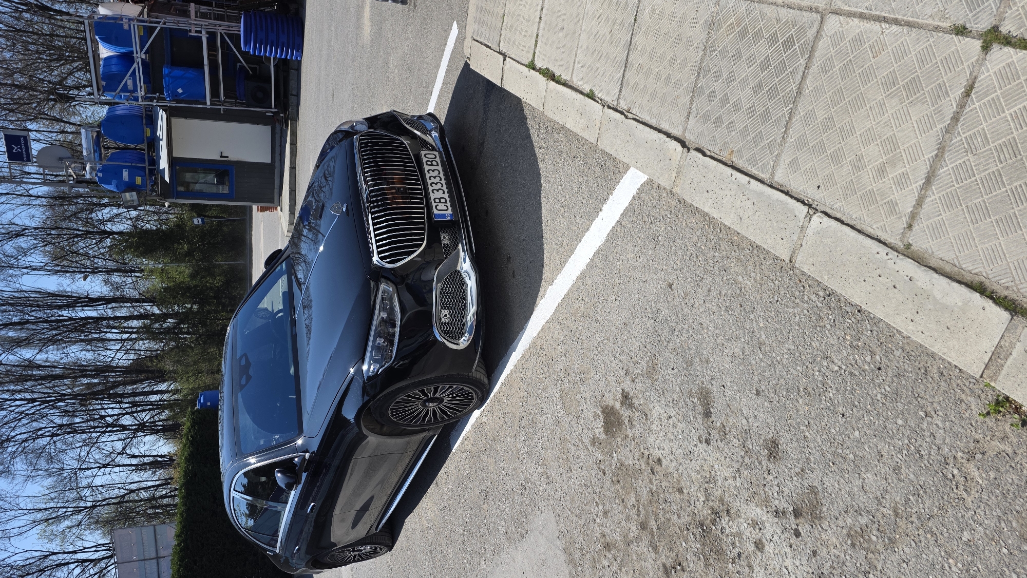 A black luxury car, identified as a Mercedes-Benz Maybach S-Class by its distinctive grille and hood ornament, is parked at an angle in an outdoor lot on a sunny day. The car occupies a space marked by white lines on asphalt pavement. Its license plate, "CB 3333 BO", is visible on the front, indicating a Bulgarian registration.
To the left and rear of the car, a structure with multiple blue barrels stacked on metal scaffolding is present, accompanied by a small utility building with a white door. A sign bearing the "Grundfos" logo (a stylized 'X' within a square) is attached to this structure. In the background, tall, bare-branched trees are visible against a clear blue sky, suggesting early spring or late autumn. A dense green hedge is also visible beyond the car's side.
The parking area is bordered by a concrete curb separating it from a patterned sidewalk made of square concrete slabs. Strong shadows cast by the car indicate bright daylight conditions. The scene appears to be an industrial or commercial area in Varna, Bulgaria.