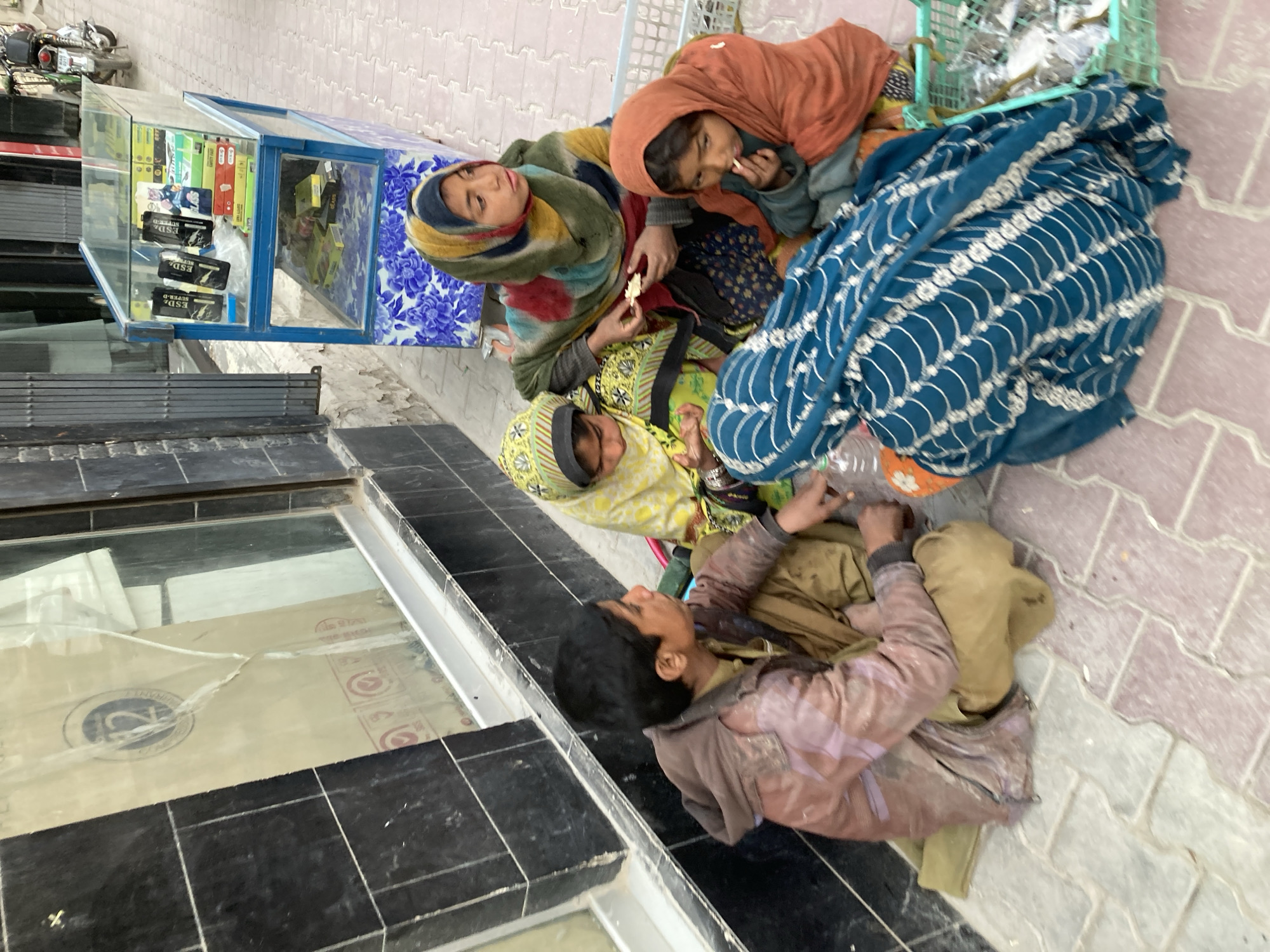 The image appears to be taken in Quetta City Tehsil, Pakistan, and depicts a group of individuals situated on the ground near a storefront.
There are four people in the image. Three of them are women wearing headscarves, while the fourth is a young boy. The women are sitting on the ground, and they seem to be eating. There is also a blue patterned blanket near them.
The young boy sits near the women. He wears a brown jacket and light brown pants.
Beside them is a storefront, with glass windows and displays. The storefront appears to be a small shop selling various items, including what looks like phone cases and electronic accessories. A motorbike can be seen parked up the street.
