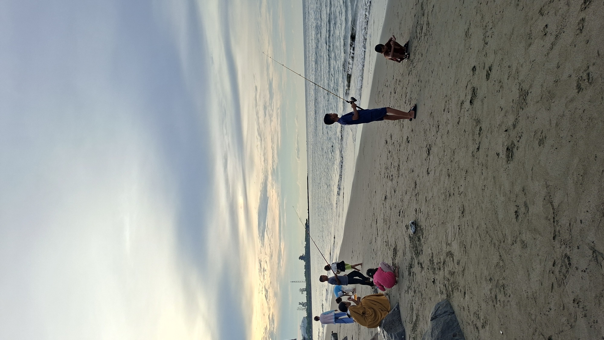 The image captures a serene beach scene in Lhokseumawe, Indonesia, likely during the late afternoon or early evening, given the soft lighting and diffused sky. Several individuals, predominantly children and a few adults, are present on the sandy shore. In the foreground, a young boy stands with a fishing rod, actively engaged in fishing, while another young boy crouches nearby on the sand, seemingly occupied with something on the ground. Further to the left, a group of people are gathered. One person, dressed in a yellow garment, is seated. Two children are also visible in this group, one kneeling and the other standing. An adult in a white shirt and blue pants is also present. In the background, the gentle waves of the ocean meet the beach, and a faint horizon line is visible with some distant structures. The sky above is a blend of soft grays and pale yellow, suggesting either an approaching storm or a fading sunset. The overall atmosphere is peaceful and recreational, depicting a common coastal pastime.