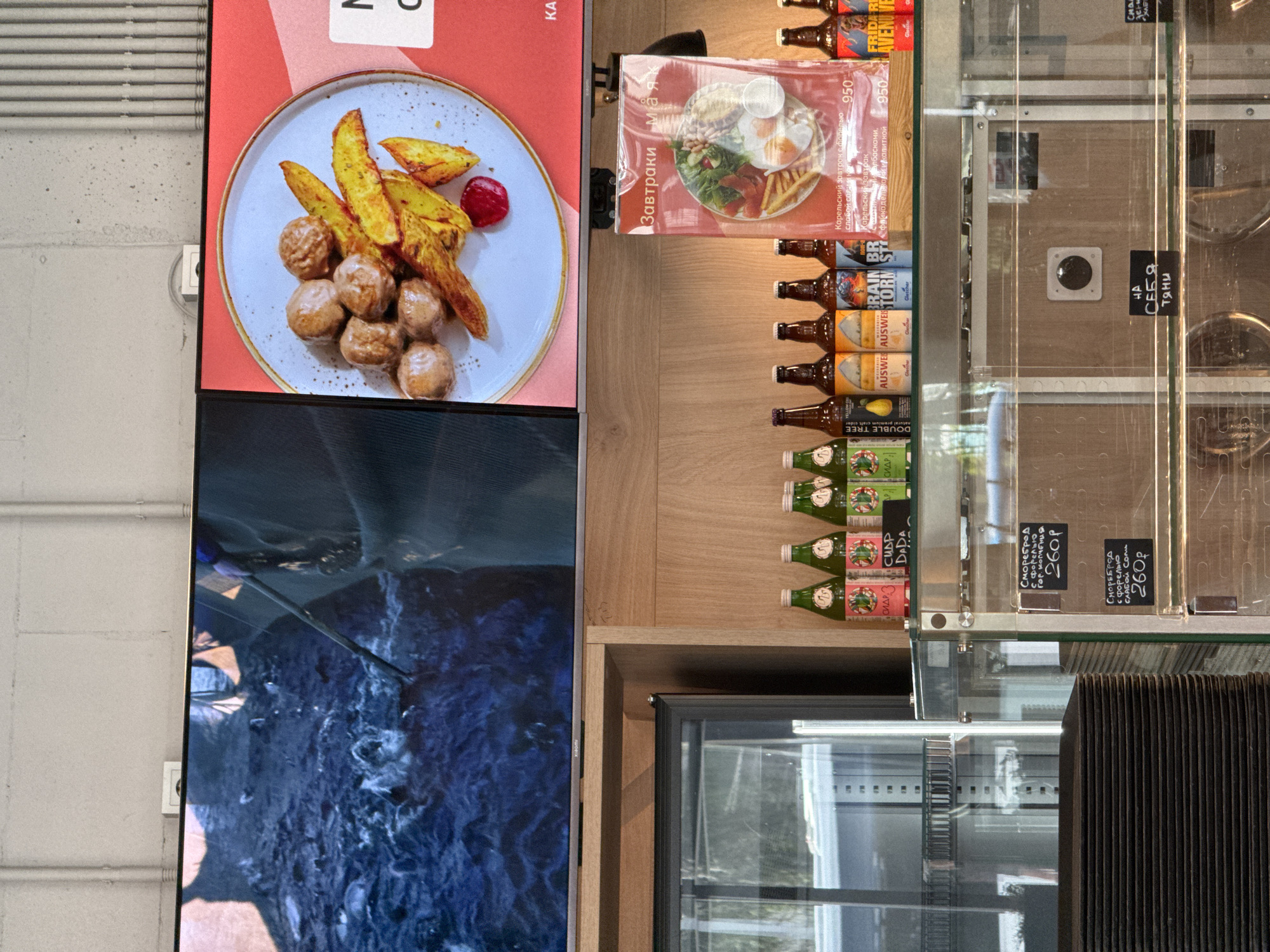 The image shows a retail display case filled with various bottled drinks and food items. On the top shelf, there are several bottles of what appears to be cider or other beverages. Below this, a glass display case reveals stacked black plates. Further down, there are chalkboards with handwritten prices and descriptions of food items. Behind the display case, a wooden wall is visible, with promotional posters for food items. The overall environment suggests a cafe or restaurant setting. The image does not provide any specific clues about the city of Lakhdenpokhya, Russia.