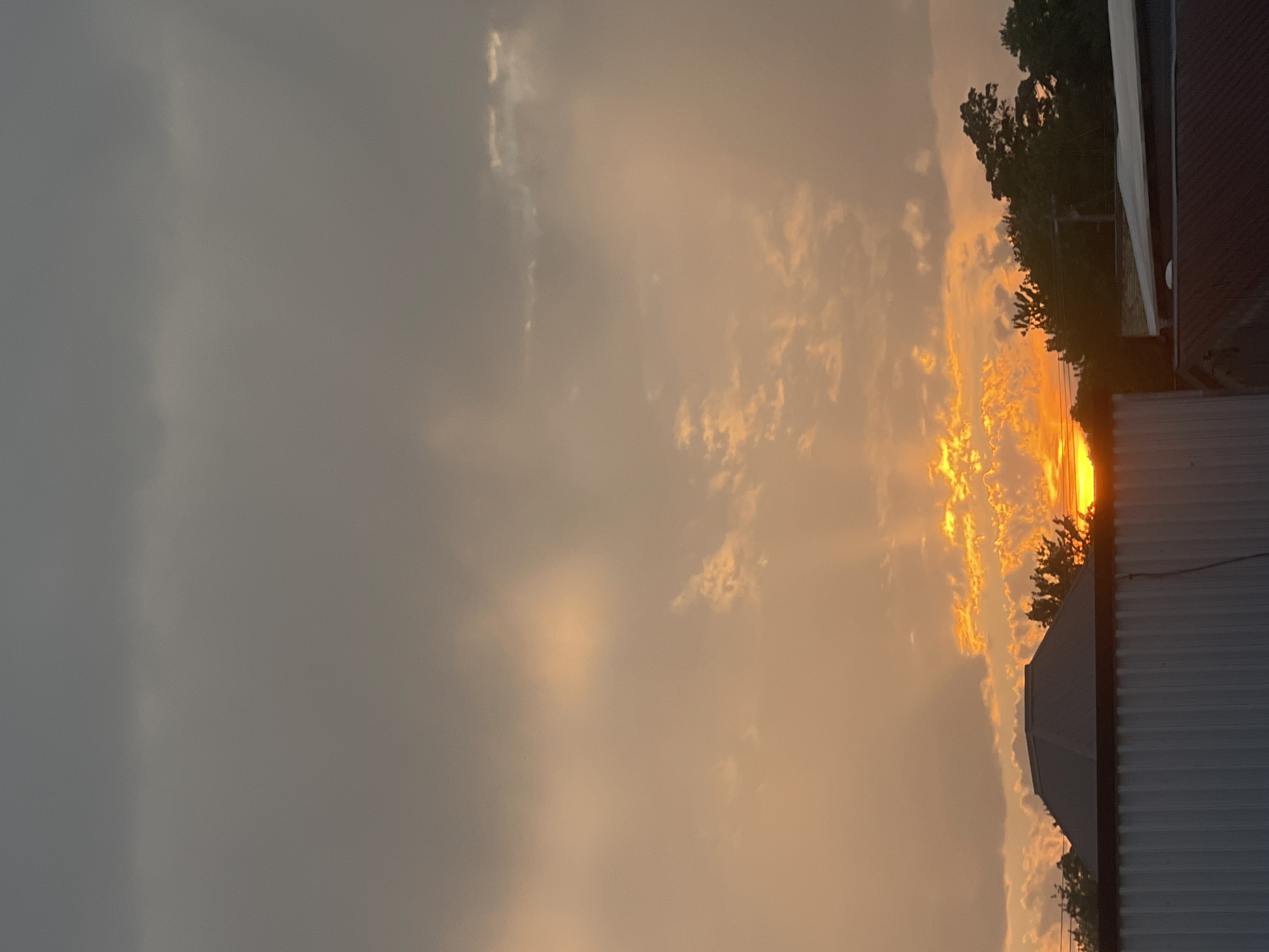 A sunset is visible over Bluewater, Canada. The sky is filled with a mix of dark grey and orange clouds. Rays of sunlight beam through the clouds, illuminating patches of sky and casting light on the tops of trees. In the foreground, the roofs of several buildings are visible, including one with a dark, multi-sided roof. The image appears to have been taken from a slightly elevated perspective, looking towards the horizon.