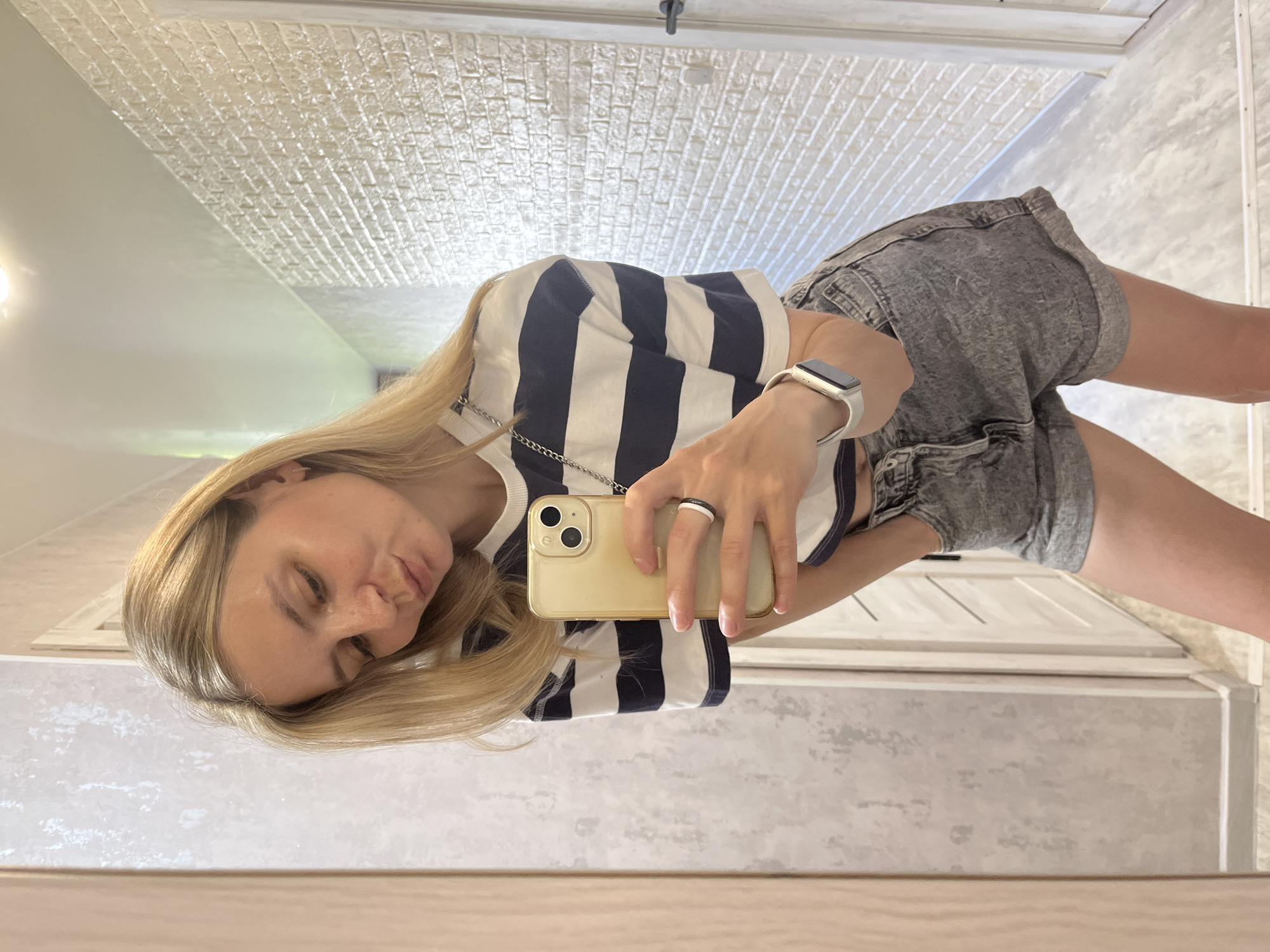 A woman is taking a selfie in what appears to be a bathroom or hallway. She has blonde hair and is wearing a striped shirt and grey shorts. She is holding a gold iPhone in her right hand. The background shows a tiled ceiling and a white door or cabinet. The location is in the city of Chaikovsky, Russia.