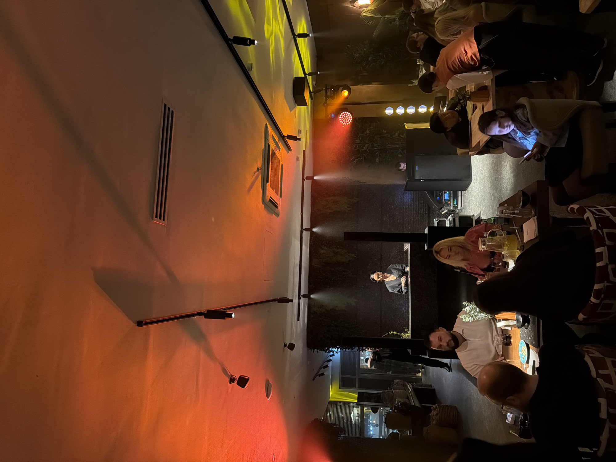Here is a description of the image:
The image shows an outdoor dining area in Moscow, Russia, at night. The area is illuminated with decorative lighting, including spotlights and colored lights on a nearby wall. People are seated at tables, eating, drinking, and socializing. One person is looking at their phone. A DJ booth is set up with a DJ performing, indicated by the person standing and equipment. Mist or smoke is present, adding to the atmospheric effect. The environment appears to be a restaurant or lounge with both indoor and outdoor seating.