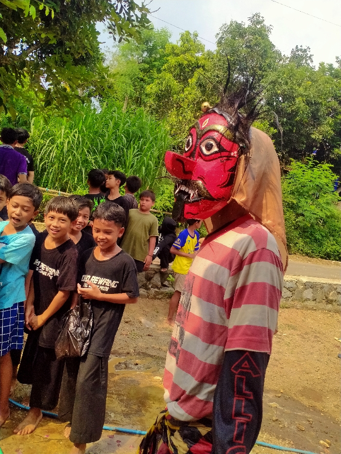 This image captures an outdoor scene in Gebyog, Indonesia, featuring a masked individual and a gathering of approximately ten children. In the right half of the frame, a person is depicted wearing a large, elaborate mask that is predominantly red and gold. The mask features a prominent red nose, white fangs, a black tongue-like appendage, and wide white eyes with black pupils, accented with gold details and surmounted by black, spiky elements. A light brown fabric extends from the back of the mask, covering the head. The masked individual wears a red and white horizontally striped long-sleeve shirt; the visible left sleeve is black with vertical red lettering "ALLIG". A yellow, black, and red patterned fabric is visible around the waist. The figure is positioned facing towards the right.
To the left and slightly behind the masked figure, a group of young males, primarily children, are gathered, observing the scene. Several children are smiling; one in the immediate foreground wears a light blue t-shirt and plaid shorts. Two other boys in the mid-ground wear black t-shirts, both displaying the text "GO REWANG", with one holding a black plastic bag. Some children in the group are barefoot.
The background is dominated by lush green vegetation, including tall, dense light green grasses or reeds in the mid-ground and darker green trees with dense canopies. The sky is partially visible, appearing light blue. The ground in the foreground is a yellowish-brown dirt path with visible wet areas, and a blue hose is present on the ground in the lower-left. A light-colored stone structure and an upward-sloping dirt path are visible in the background on the right.
