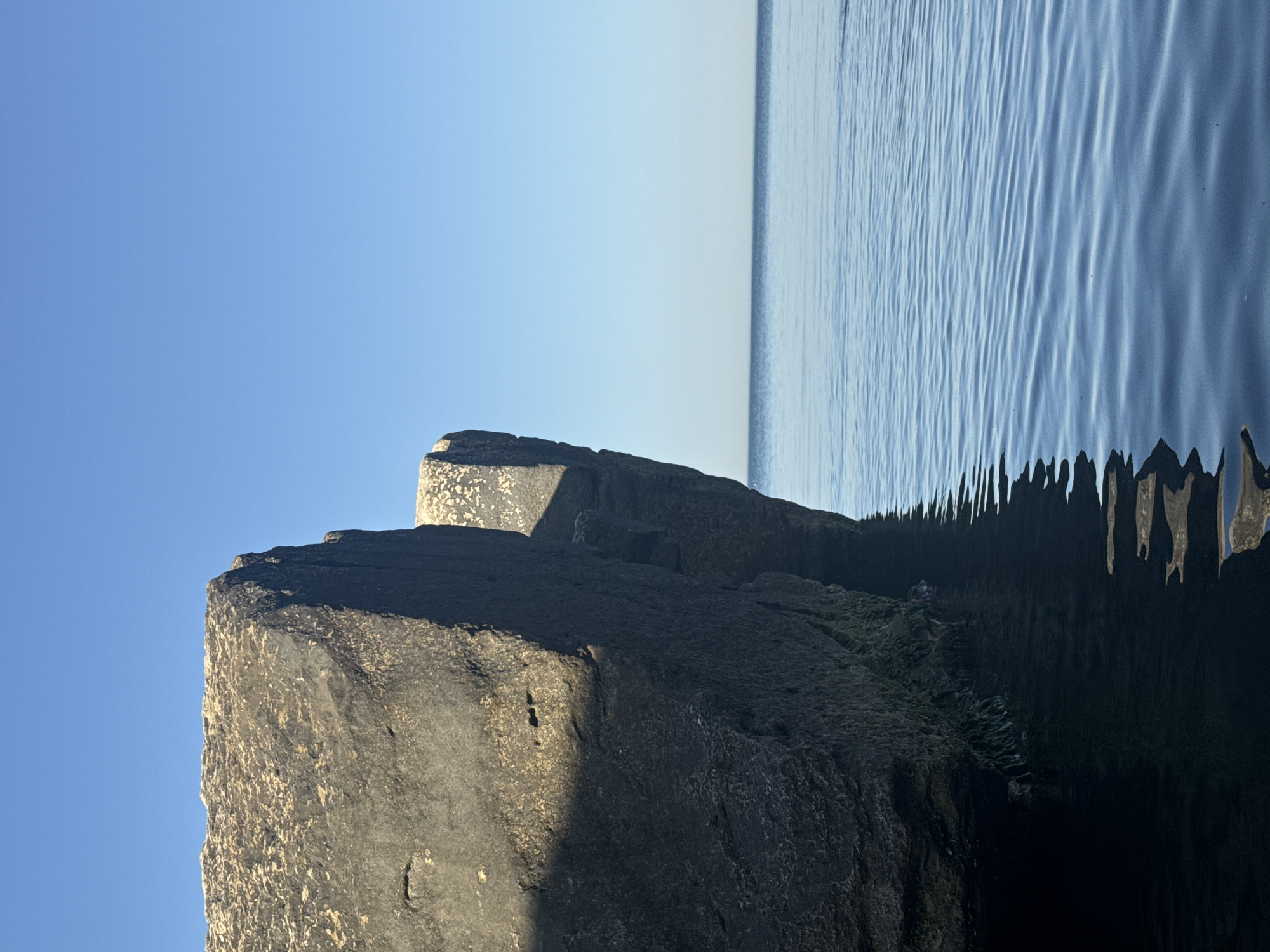 The image shows large, dark gray rocks emerging from the calm, blue waters of the Caspian Sea. The sky is a clear, bright blue. The rocks appear to be part of a man-made structure, possibly a breakwater or pier, located in the city of Aktau, Kazakhstan. The water is shallow around the base of the rocks, with some green algae visible clinging to their submerged surfaces. The light suggests it is daytime, with the sun casting shadows on the rocks. No people or other objects are visible in the frame.