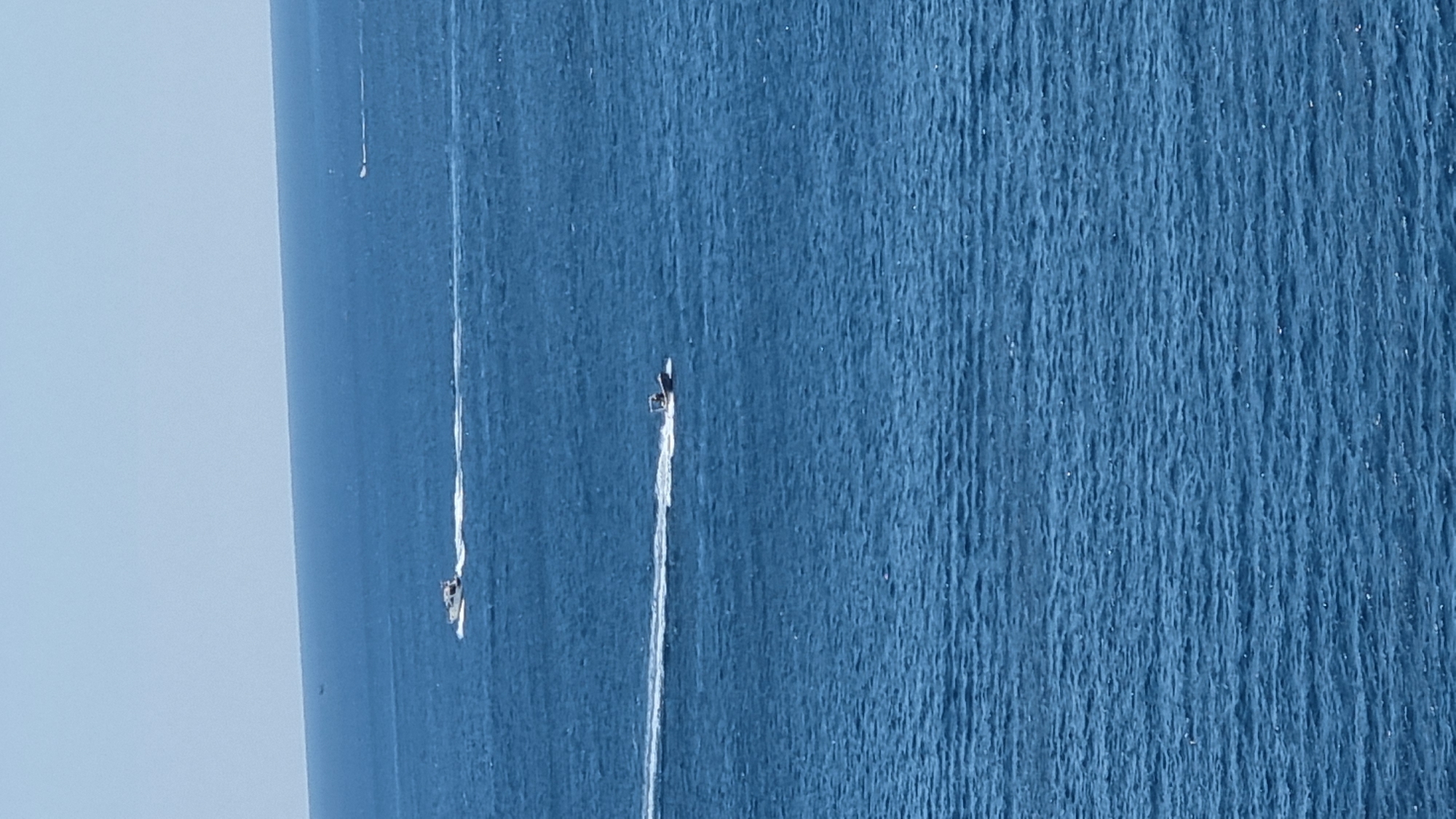 The image depicts a vast expanse of deep blue ocean under a clear, pale sky. The horizon line is clearly visible, separating the water from the sky. Two boats are actively moving across the water, each leaving a distinct white wake behind them. The boat in the foreground is closer and larger in the frame, appearing to be a motorboat with a dark hull and a small cabin or canopy. The second boat is further in the distance, smaller and less distinct, also leaving a trail of white spray. The overall impression is one of an open sea on a bright day, with activity on the water. The location is identified as the sea off the coast of Roses, Spain.