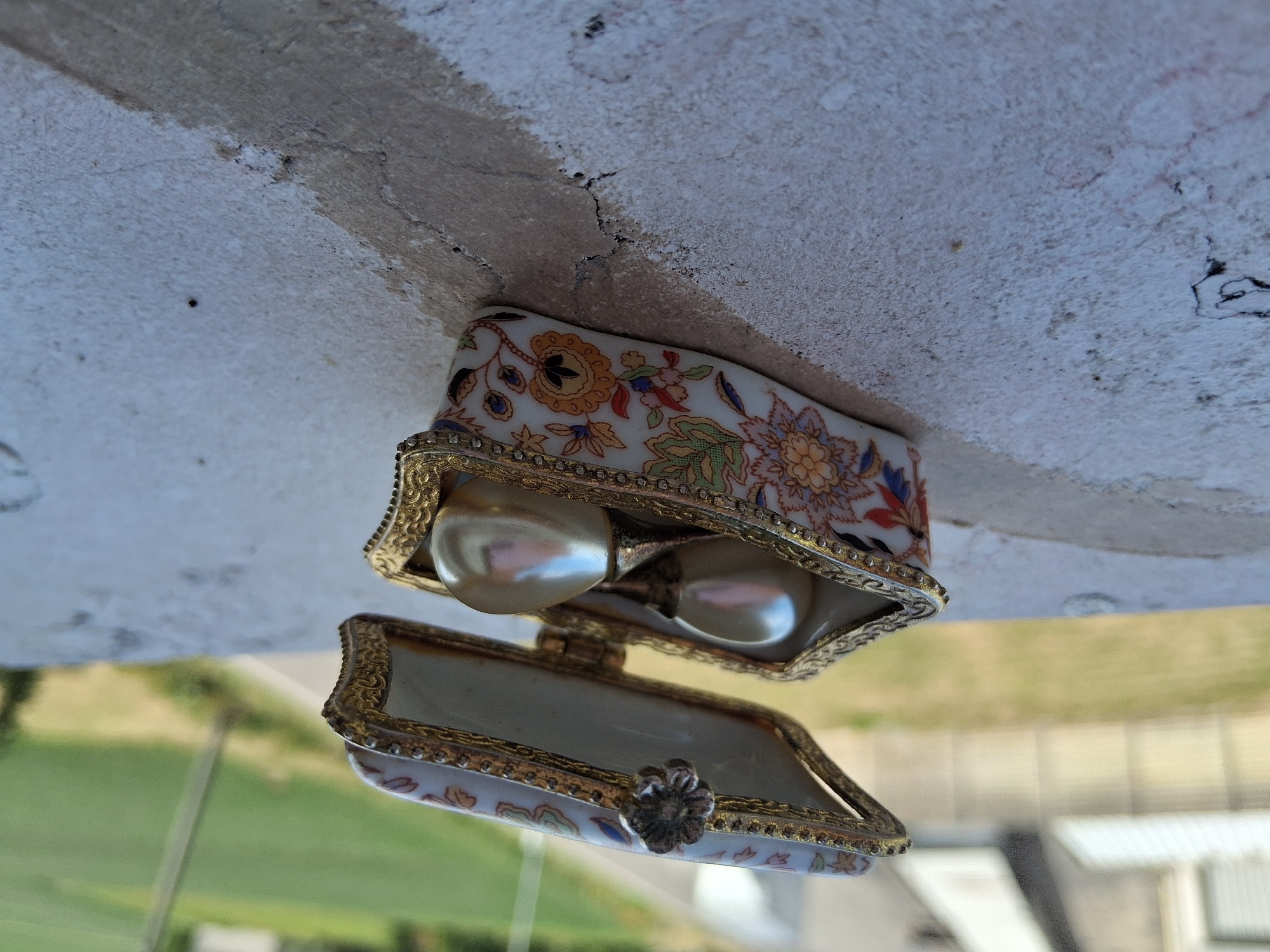 A small, ornate porcelain jewelry box with a floral pattern and gold trim sits on a textured concrete surface. The lid of the box is open, revealing two pearlescent decorative balls inside. The background is blurred, showing a grassy area and what appears to be a building with a corrugated metal roof. The scene suggests an outdoor setting, possibly a balcony or ledge, in the city of Colorno, Italy, given the context.
