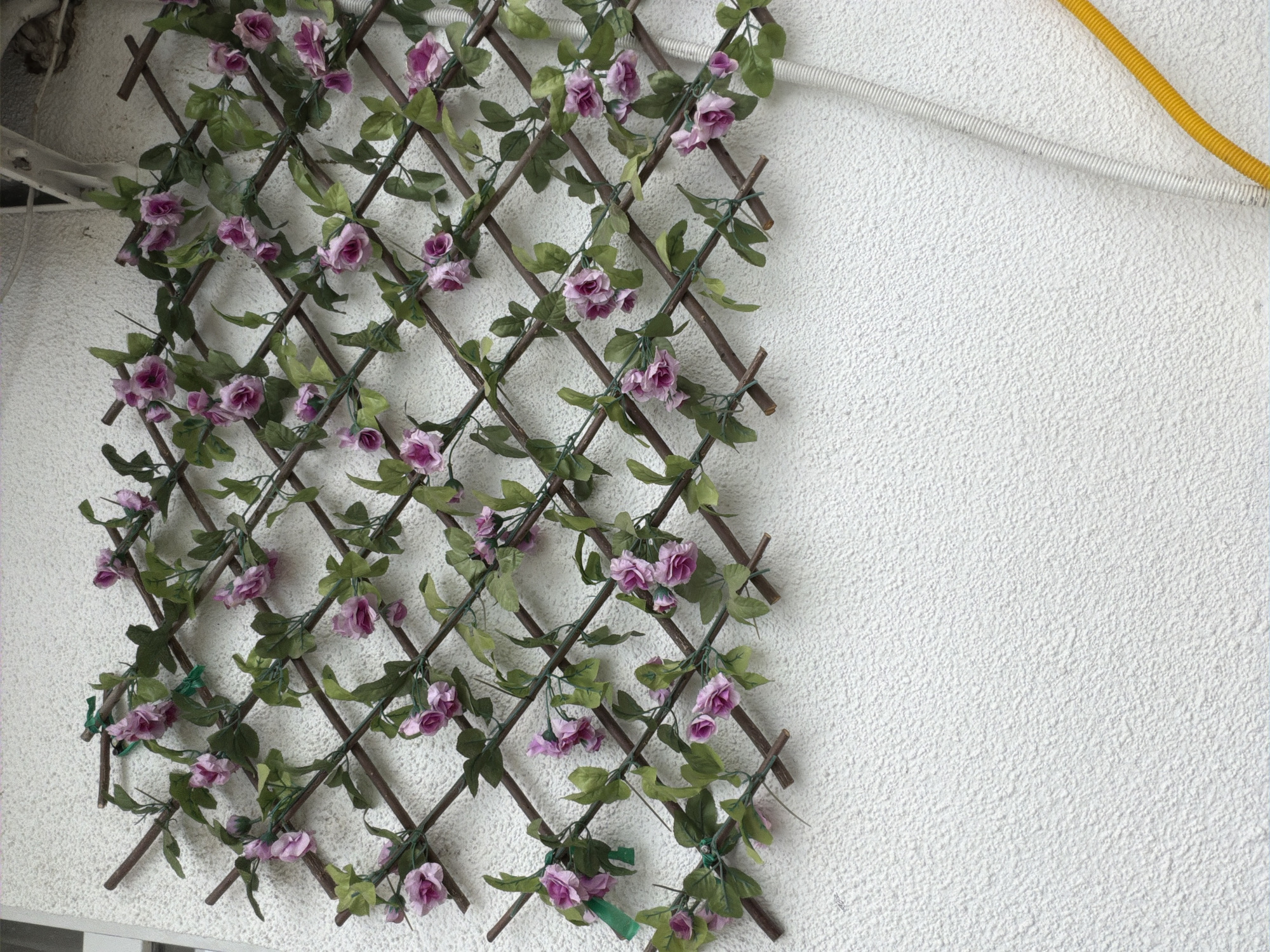 The image shows a section of a wooden lattice covered with artificial purple roses and green leaves, mounted on a textured white wall. The lattice is arranged in a diamond pattern, with the vines and flowers woven through it. To the upper right of the lattice, two cables are visible against the wall: one is a white, ribbed cable, and the other is a bright yellow, ribbed cable. The scene appears to be outdoors, as indicated by the sunlight on the wall and the presence of exterior cables. The overall impression is one of decorative outdoor furnishing or perhaps part of a balcony railing. The location context of Tbilisi, Georgia, suggests this might be a typical sight on a balcony or garden wall in the city. There is no text visible in the image.