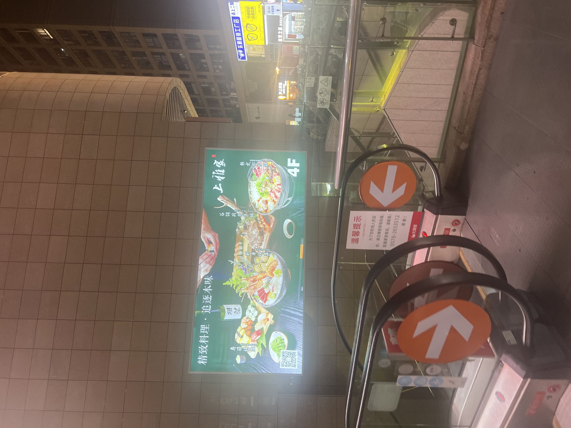 This image was taken at night in Liandu District, China. The foreground shows the top of an escalator with orange circular signs displaying white arrows pointing left and right. Behind the escalator, a large illuminated billboard displays images of Japanese cuisine, including sushi, sashimi, and a hot pot. Text in Chinese is visible on the billboard, with the words "精致料理·追本味" (Exquisite cuisine, trace the original flavor) and "上雅家" (Shang Yajia) prominently featured. The billboard also shows a QR code and the text "4F", indicating the floor number. In the background, a tall building with numerous lit windows is visible, along with other businesses and signs. A "溫馨提示" (Warm reminder) sign is also visible on the escalator, cautioning against using the escalator while holding children or pets.