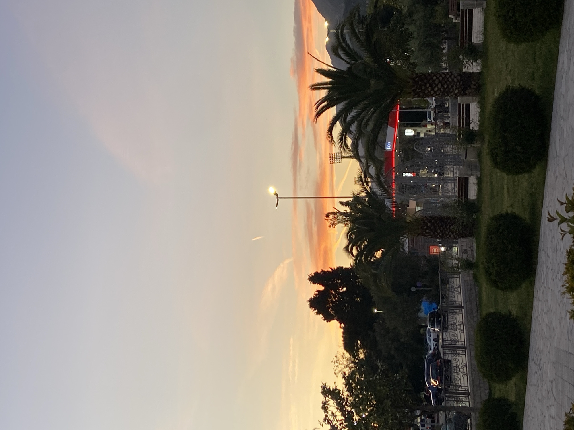 The image captures a serene urban scene at sunset or dusk in Bar, Montenegro. The sky dominates the upper portion, transitioning from a pale blue-grey on the left to warm hues of orange and pink on the right, indicating the setting sun. A faint, white airplane contrail stretches diagonally across the lighter part of the sky.
In the mid-ground, a single bright streetlamp stands illuminated, casting a glow. Several tall palm trees with dark, textured trunks and prominent fronds are scattered across the lower half of the image, silhouetted against the warm sky. In the background on the right, parts of a mountain range are visible, bathed in the fading light.
At street level, on the right, a building with a large red sign is visible, displaying the text "EKO," likely a gas station or a store. Other structures and lights are faintly discernible behind the palm trees and the fence. An ornate black metal fence, interspersed with white decorative elements, runs along what appears to be a sidewalk or median strip. Beyond the fence, well-maintained green bushes and grassy patches line the area. A few cars are visible on the street, some parked and others possibly in motion. One person, wearing a blue shirt, can be faintly seen near a vehicle. The overall atmosphere is tranquil and peaceful, showcasing the quiet transition of day to evening in a coastal town.