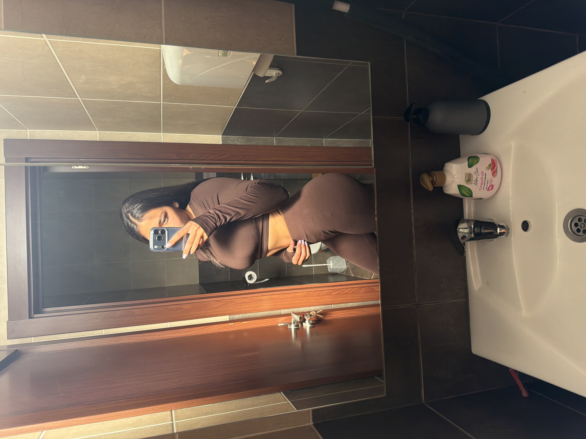 The image shows a person taking a mirror selfie in a bathroom. The bathroom has a white sink with a faucet, and two bottles of soap are visible on the counter next to the sink. The bathroom walls and floor are tiled with a mix of light brown and dark brown tiles. A wooden door is partially visible in the reflection of the mirror. The person taking the selfie is wearing a brown, long-sleeved outfit. There is no information in the image to connect it to the city of Gabrovo, Bulgaria.