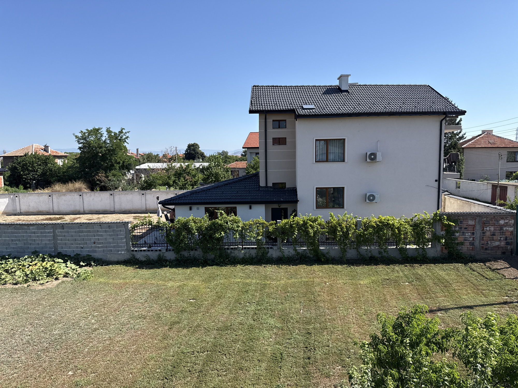 In Dragor, Bulgaria, on a clear, sunny day, a two-story white house with a dark tiled roof stands in a suburban setting. The house has several windows with brown frames and curtains, and two outdoor air conditioning units are mounted on its facade. Adjacent to the house is a smaller section with a dark tiled roof and a light-colored wall with windows. A fence covered in green vines separates the property from the front yard, which features a well-maintained lawn and a patch of what appears to be a vegetable garden. In the background, other houses with red tiled roofs and trees are visible under a bright blue sky. The scene suggests a peaceful residential area.