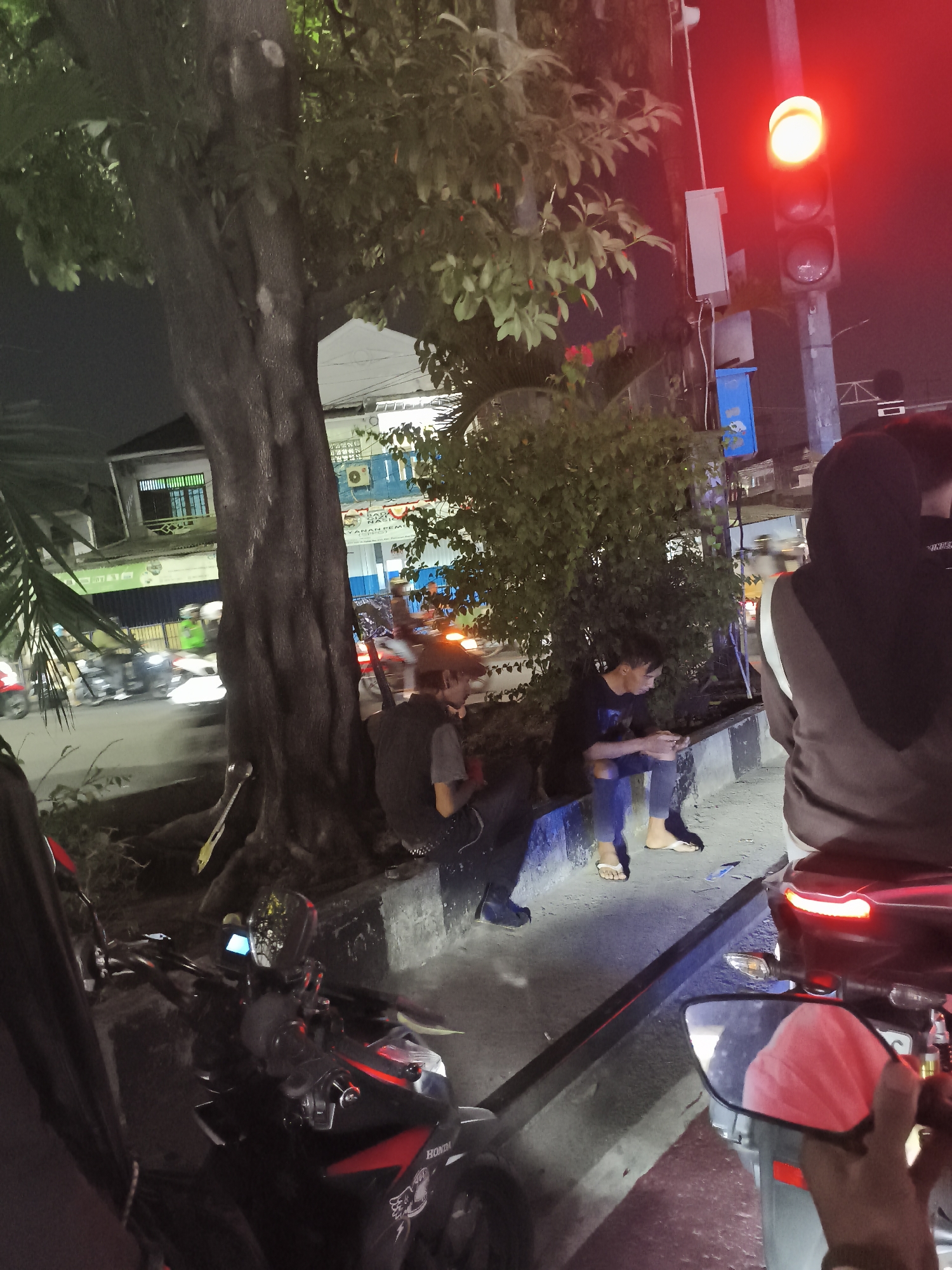In Depok, Indonesia, a street scene at night unfolds near a traffic light. Two individuals are seated on a low wall beside a large tree. One, wearing a dark vest over a grey t-shirt and dark pants, is looking down. The other, a younger male in a black t-shirt and ripped blue jeans, is holding a mobile phone and also looking downwards. In the foreground, a motorcycle is parked, its red taillight illuminated, and a mirror reflects a person wearing a pink head covering. Other motorcycles and some buildings are visible in the background, suggesting a busy urban environment. The traffic light is red, indicating a stop in traffic.