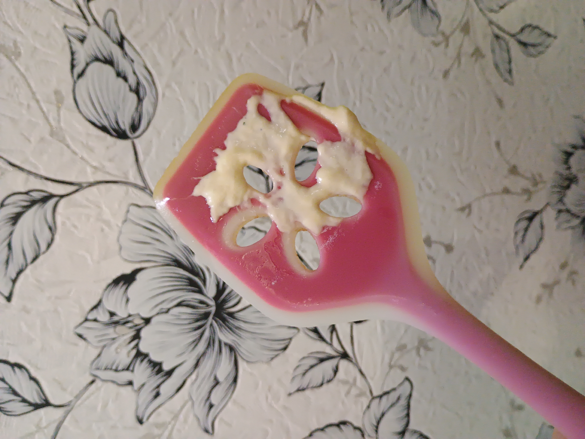 A pink and white silicone spatula is held up in front of a textured wallpaper with a floral pattern. The spatula head has cutouts and is partially covered in a creamy, light-colored substance, possibly food. The wallpaper depicts stylized black and white flowers and leaves. The scene is indoors. The image does not contain information about the city of Zaporizhzhia, Ukraine.