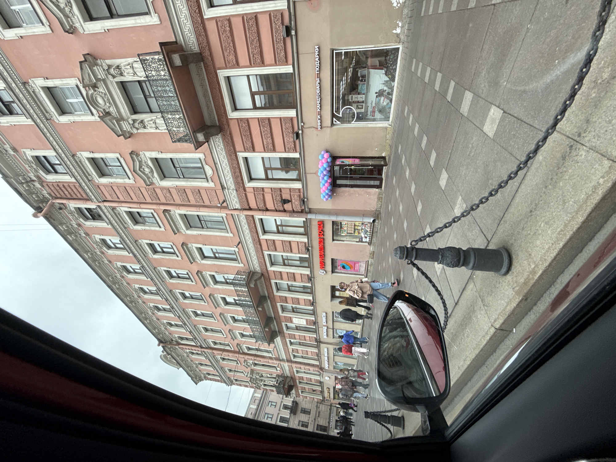 This image captures a street scene through the window of a vehicle, featuring architecture and pedestrian activity. The perspective is angled upwards towards a tall, ornate building with a pink facade, multiple stories, and numerous windows, some of which have small balconies.
A store front is visible with the text "КНИГИ КАНЦТОВАРЫ ПОДАРКИ" vertically written on the building. Another store front is visible on the building with the text "КОНДИТЕРСКИЙ МАГАЗИН ШОКОЛАД КОНФЕТЫ". The storefront has a decorative balloon arch in blue and pink.
On the sidewalk, a small group of people are walking. A chain fence with black posts runs along the edge of the sidewalk, closer to the viewpoint. The side mirror of the vehicle partially obscures the view of the sidewalk.
Other text visible includes "ОМАРМЕЛАДНАЯ БОЧКА" and "INCANTO", possibly indicating store names. The OCR also detected "буквоед".
Based on the architecture and the Cyrillic script, the location is likely in Russia, though the specific city is not identifiable from the image alone.