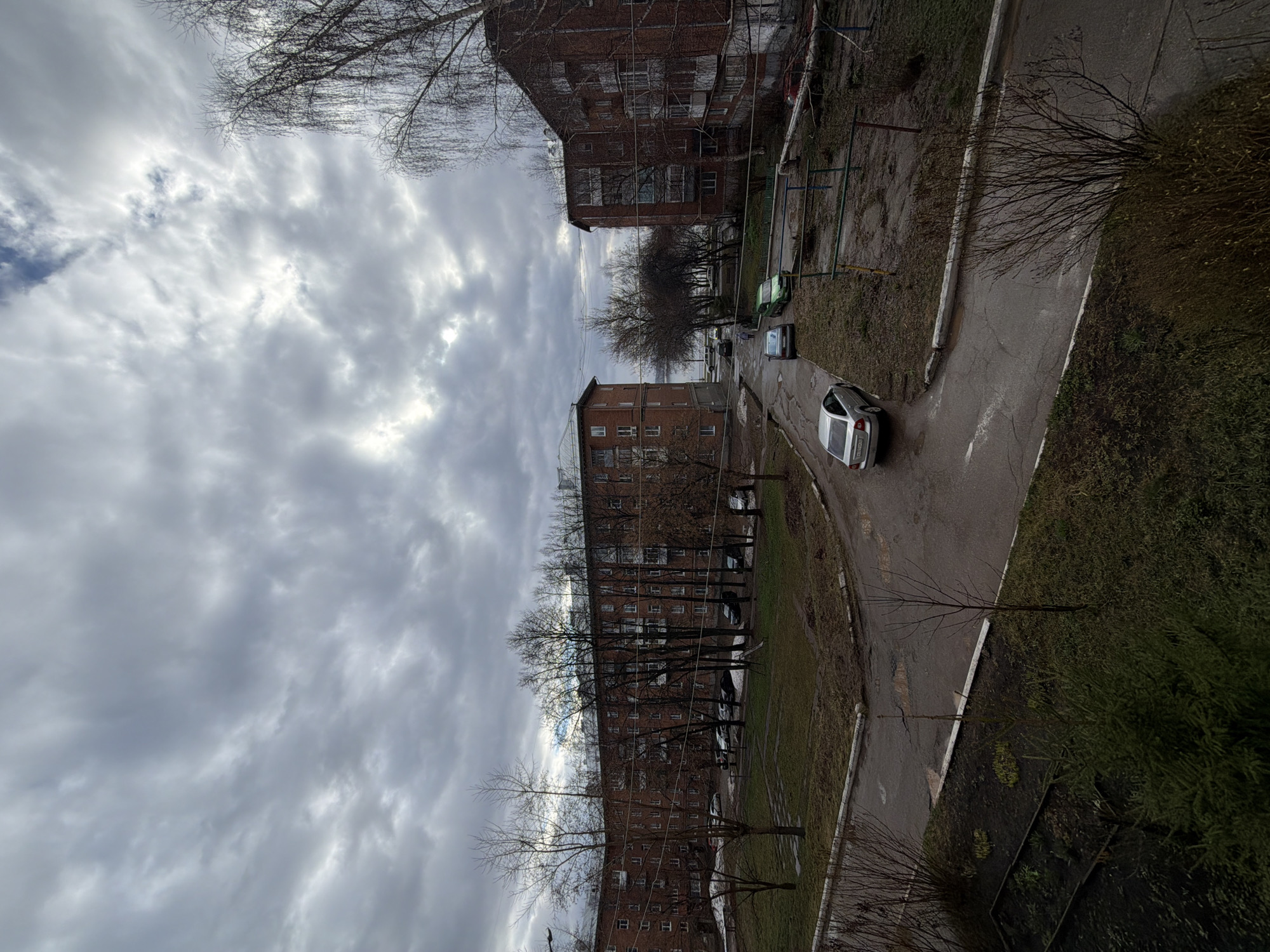 This elevated photograph captures a residential area in Votkinsk, Russia, under an overcast sky, characteristic of late autumn, winter, or early spring. The scene is dominated by several multi-story brick apartment buildings, typical of Soviet-era architecture, featuring numerous windows and fire escapes.
A winding, damp asphalt road with white painted lines curves through the middle ground, indicating recent rain or snowmelt. Several cars are visible on or alongside the road; a silver sedan is prominent in the mid-right, with a partially obscured license plate. A green car is parked further down, and other vehicles are scattered among the leafless deciduous trees that line the road and stand between the buildings. The ground consists of patches of dry, sparse grass and bare shrubbery.
The sky is heavily laden with dense, grey clouds, contributing to a muted and subdued atmosphere, though brighter patches suggest the sun is behind the cloud cover. Faint utility lines are visible strung between buildings, and some metal structures, possibly playground equipment or utility poles, can be seen near the road. The overall impression is one of a quiet, utilitarian urban landscape on a cloudy day, with no specific human activity or event clearly discernible beyond the presence of vehicles. No distinct text is clearly legible.