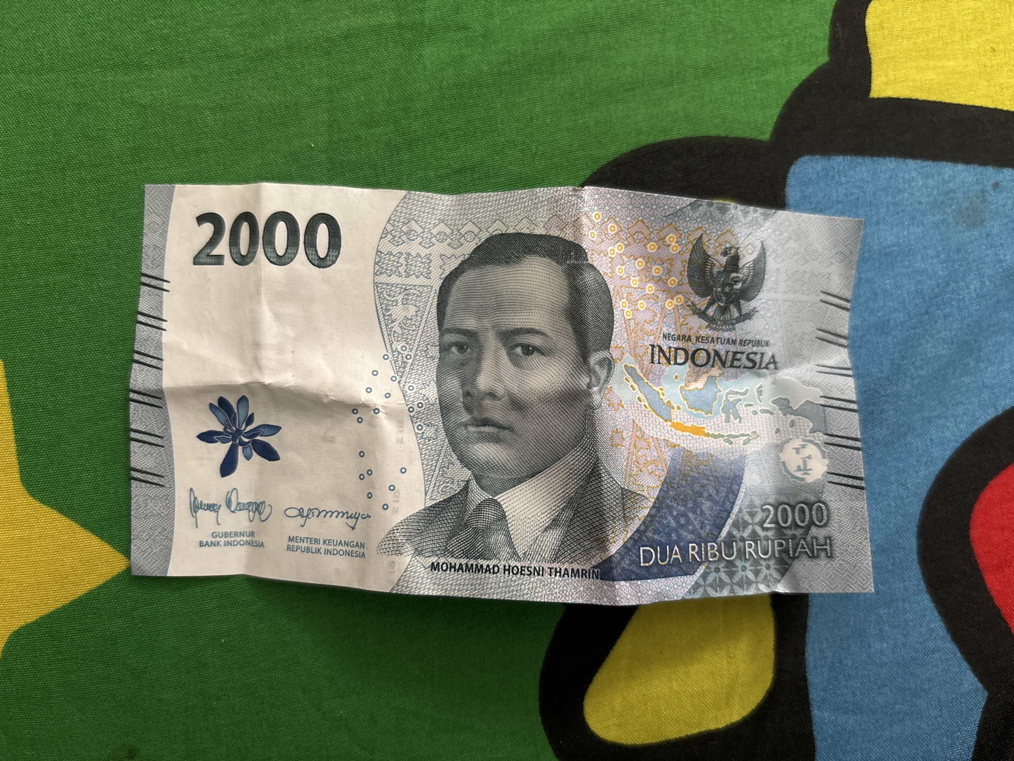 A slightly crumpled 2000 Indonesian Rupiah banknote is displayed on a patterned fabric surface featuring a green background with irregular, cartoon-like shapes in yellow, black, blue, and red. The banknote depicts Mohammad Hoesni Thamrin, an Indonesian national hero, in portrait. The upper-left side of the note displays the denomination "2000". A blue flower-like design is located near signatures and official titles from Bank Indonesia. On the right side, the Garuda Pancasila, the national emblem of Indonesia, is featured, alongside a map of the Indonesian archipelago and the text "NEGARA KESATUAN REPUBLIK INDONESIA". The denomination "2000" and "DUA RIBU RUPIAH" (Two Thousand Rupiah) are shown at the bottom-right corner of the banknote. There is no indication of the image being taken in North Jakarta or any specific location.