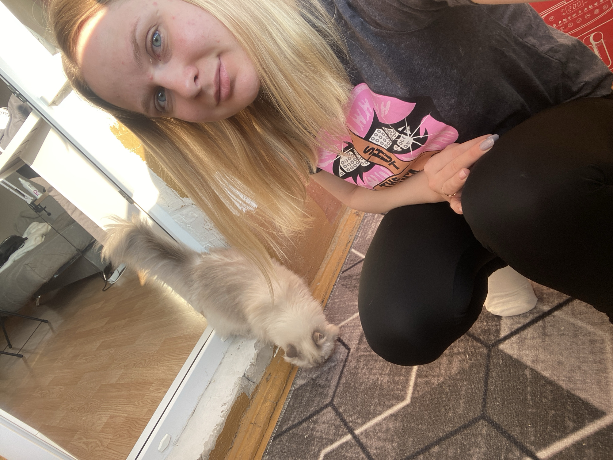 In a room, a fair-skinned woman with blonde hair and blue eyes is taking a selfie. She is wearing a dark gray t-shirt with a pink graphic and black leggings. Her left hand, with light blue nail polish, rests on her knee, adorned with a ring. Beside her, a fluffy, light-colored cat with blue eyes is sniffing the floor. The environment features a wooden floor visible through a reflective surface, a patterned rug, and a white baseboard. The lighting suggests natural sunlight streaming into the room. The image does not contain enough information to determine the location in the city of Chaikovsky, Russia.