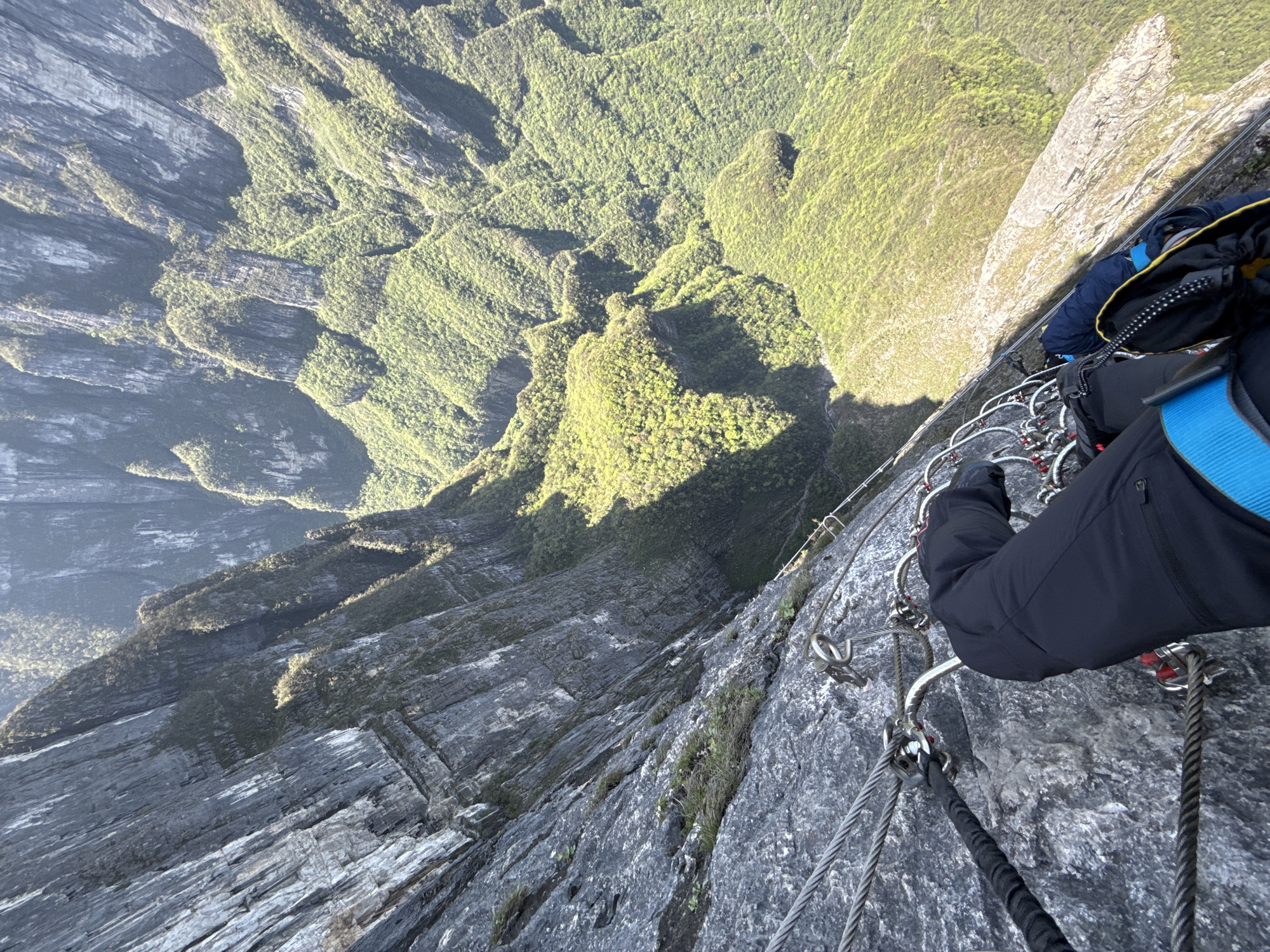 A first-person perspective shows a person secured to a via ferrata on a cliff face. The person wears dark trousers and a backpack with blue straps. Metal cables and climbing aids are fixed to the rock face. Below and to the side, a steep, green, mountainous landscape extends downwards. The location is likely Tianmen Mountain in Yongding District, China.