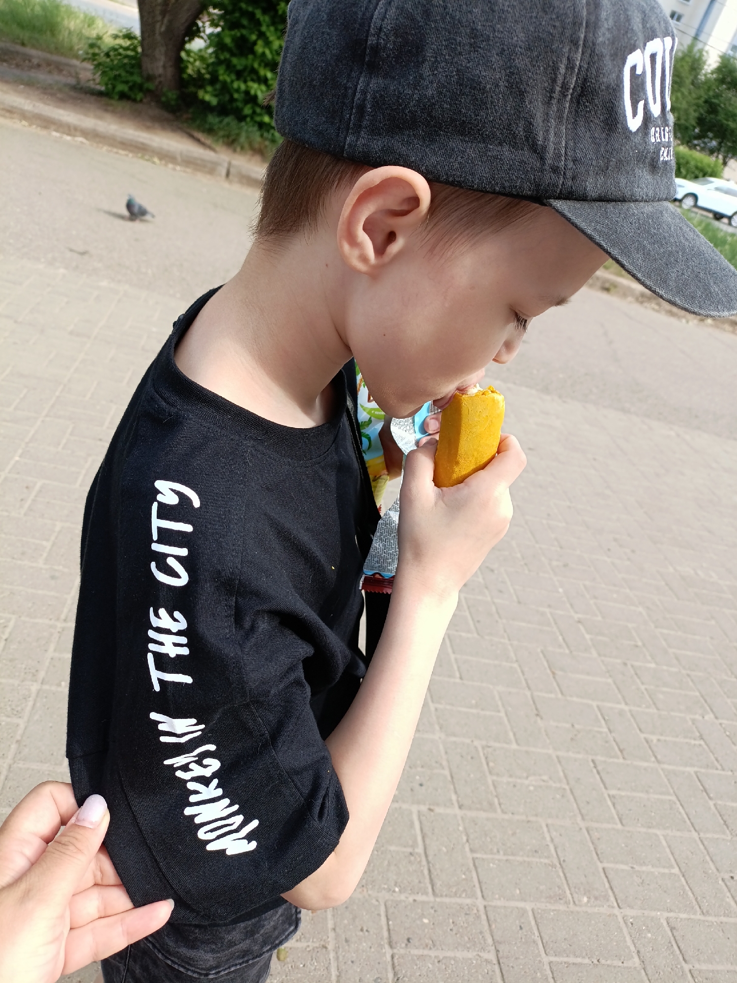 A young boy is standing outdoors, likely in Chaikovsky, Russia, based on image characteristics. He is wearing a black baseball cap and a black T-shirt with white writing on the sleeve that says "Monkey in the City." The boy is eating a yellow, packaged food item, possibly a snack. An adult hand is partially visible on the lower-left side of the image, touching the sleeve of the boy's T-shirt. The background includes a paved surface, a street, trees, a building, and a white car. A pigeon is visible in the background on the left. The scene appears to be taking place during daylight hours.