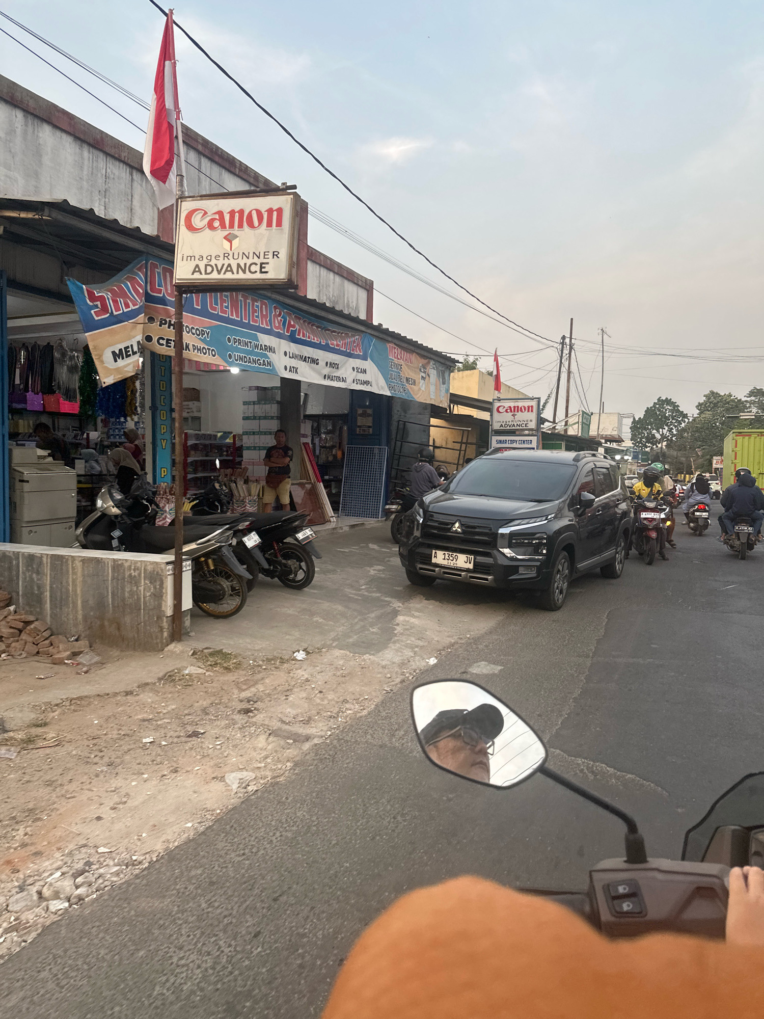 The image depicts a street scene in Saruni, Indonesia, during daylight. In the foreground, the handlebars and a portion of the seat of a motorcycle are visible, implying the photo is taken from a rider's perspective. The rider's forearm, wearing an orange sleeve, is also in view. A motorcycle side mirror reflects the rider, a person wearing glasses and a dark baseball cap, looking upwards.
The middle ground features a street with several vehicles. A dark gray Mitsubishi Xpander SUV is prominently parked on the right side of the road, displaying a license plate "A 1359 JV". Several motorcycles are also present, some parked on the side and others in motion, with riders wearing helmets. A green truck is partially visible in the far right background.
The left side of the image shows a storefront with a red and white Indonesian flag flying from a pole above a sign for "Canon imageRUNNER ADVANCE". Below this, a larger sign reads "SINAR COPY CENTER & PRINTING CENTER". The shop appears to sell office supplies and printing services, with shelves stocked with paper and other items visible inside. Two men are standing outside the shop, one near the motorcycles and another further inside. Several parked motorcycles are in front of the shop.
The environment is a street lined with buildings and a sidewalk area. Electrical wires crisscross overhead. The ground is a mixture of asphalt and dirt. The sky is overcast with diffused light. The overall impression is a busy commercial area in a developing urban setting.