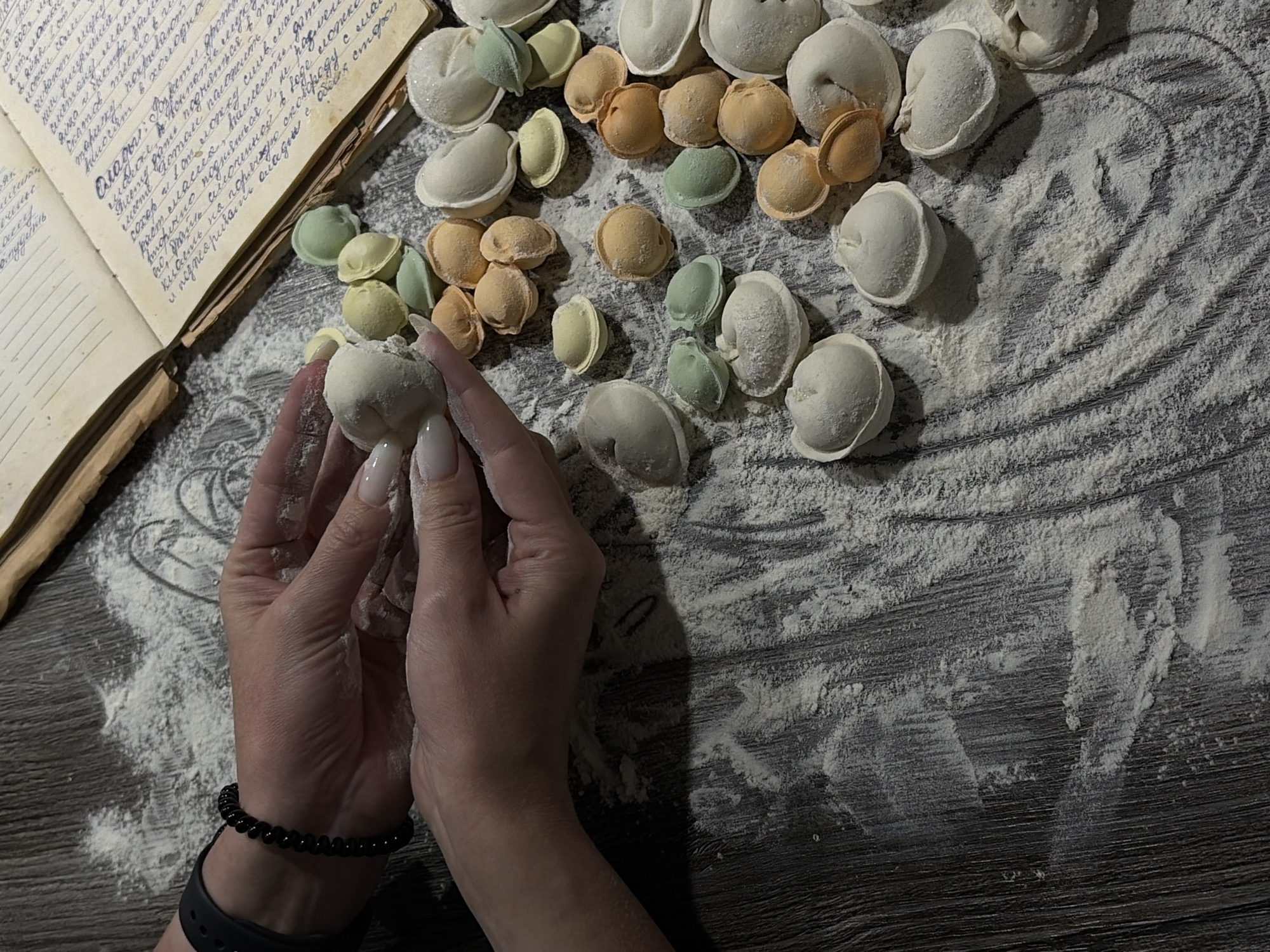 A person is preparing dumplings on a floured surface with a recipe book visible in the upper left corner. The dumplings are various colors (white, green, and orange) and different sizes. The person is holding a white dumpling between their hands. The scene is taking place in an indoor environment, likely a kitchen. The surface is a dark gray wooden table. A black beaded bracelet is on the person's left wrist along with a smartwatch band. Based on the recipe in the book, it could be assumed that the location is Chaikovsky, Russia.