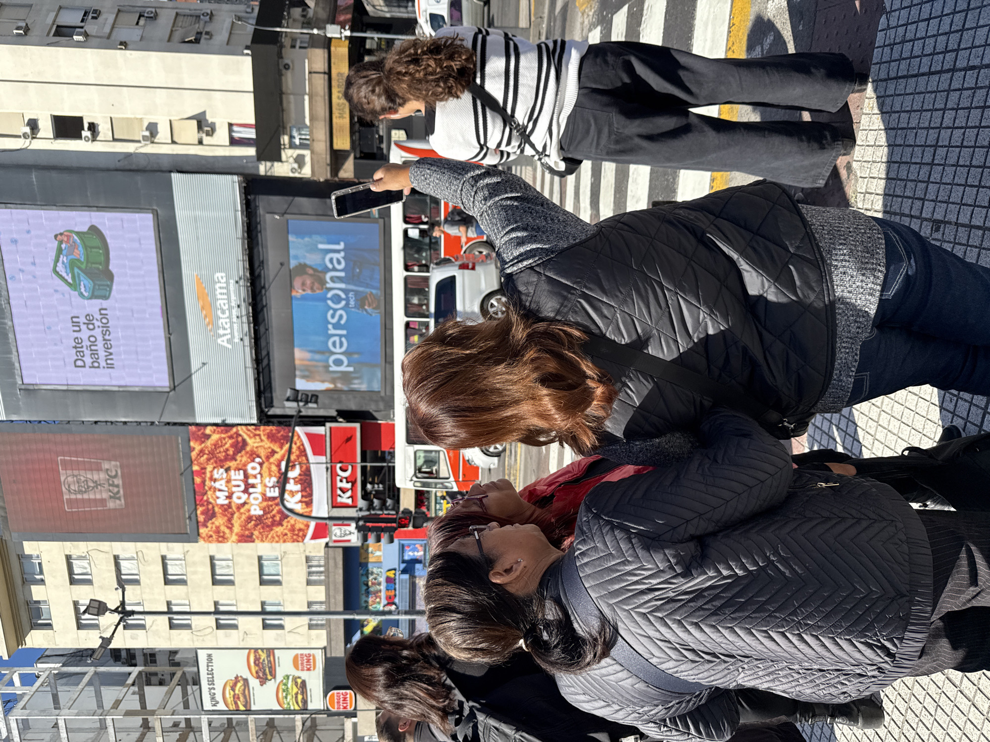 Captured from street level in the city of Buenos Aires, Argentina, the image presents a bustling urban scene. Several pedestrians are present; one is taking a photograph with a smartphone, and others are standing or moving around. In the background, urban architecture is visible, including buildings adorned with commercial advertisements. The advertisements feature brands like KFC and Burger King, as well as promotional content for companies like "personal tech" and "Atacama". The setting appears to be a busy commercial district in the Argentinian capital.