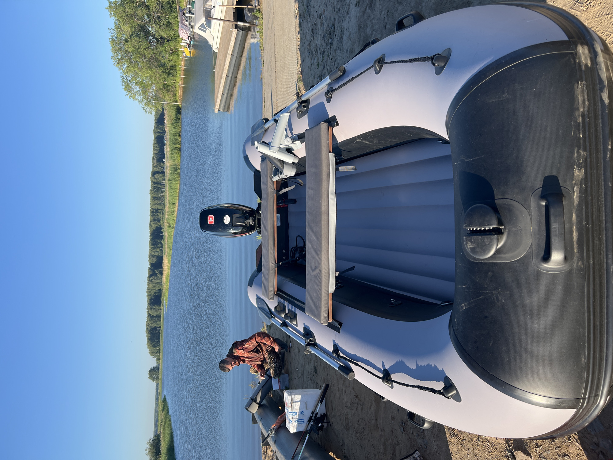 An eye-level, medium shot captures a man in a camouflage jacket and pants squatting by a gray inflatable boat on a sandy shore. The boat is equipped with a black outboard motor, two gray padded seats, and an oar mounted on its side. Fishing rods and a white cooler are placed beside another deflated inflatable boat in the background. A body of water, likely a river, stretches towards a tree-lined horizon under a clear blue sky. A wooden dock with a small white boat is visible on the right. The scene is located in Novopichugovo, Russia.