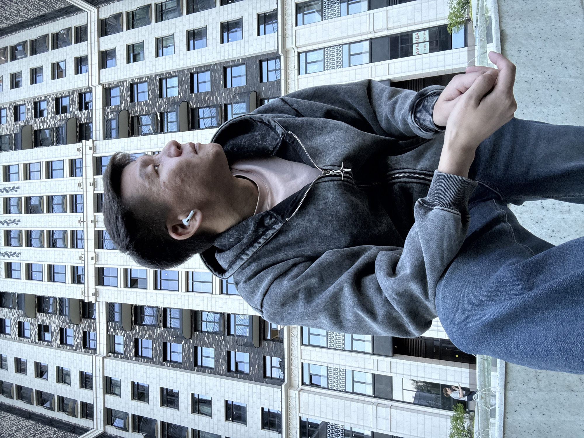 A medium shot shows a man sitting outside with a building in the background. He is wearing a gray hooded jacket over a light pink shirt, and blue jeans. He has an earbud in his left ear. His hands are clasped together. The building behind him is tall and modern, with a repetitive pattern of windows. The location is Unknown, Unknown.