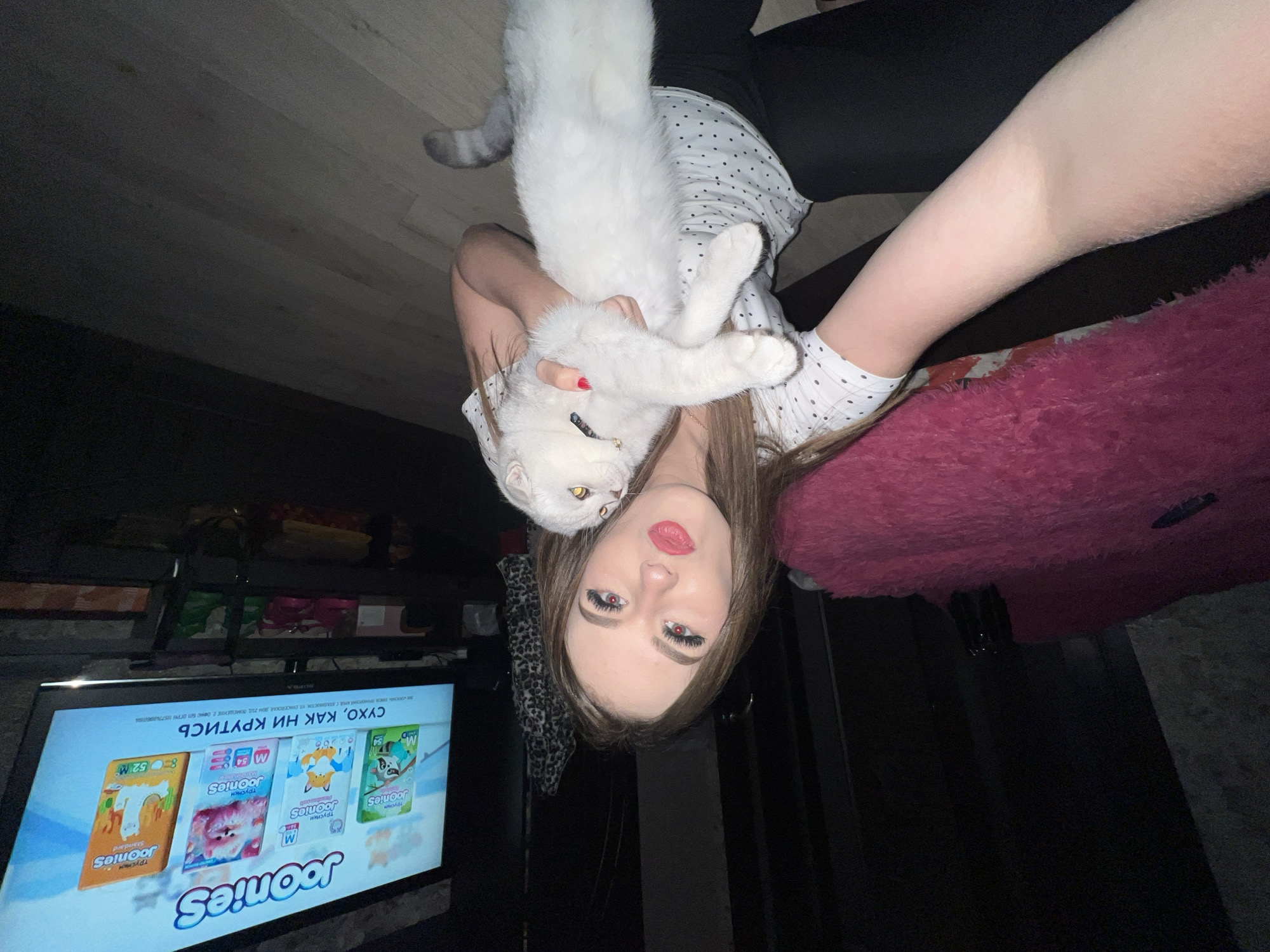 A woman is holding a white cat while lying upside down. She is wearing a white polka dot shirt and black pants. A television is in the background displaying a diaper advertisement for a brand called "Joonies." The advertisement is in Russian and includes the phrase "СУХО, КАК НИ КРУТИСЬ." The text "Г. АЛАДИВОСТОК, УЛ. ЕНИСЕЙСКАЯ, ДОМ ГОД ПОМЕЩЕНИЕ 2. ОФИС 501 ОГРН 11577469604" is also visible on the television screen. The location of Vladivostok, Russia is mentioned on the television.