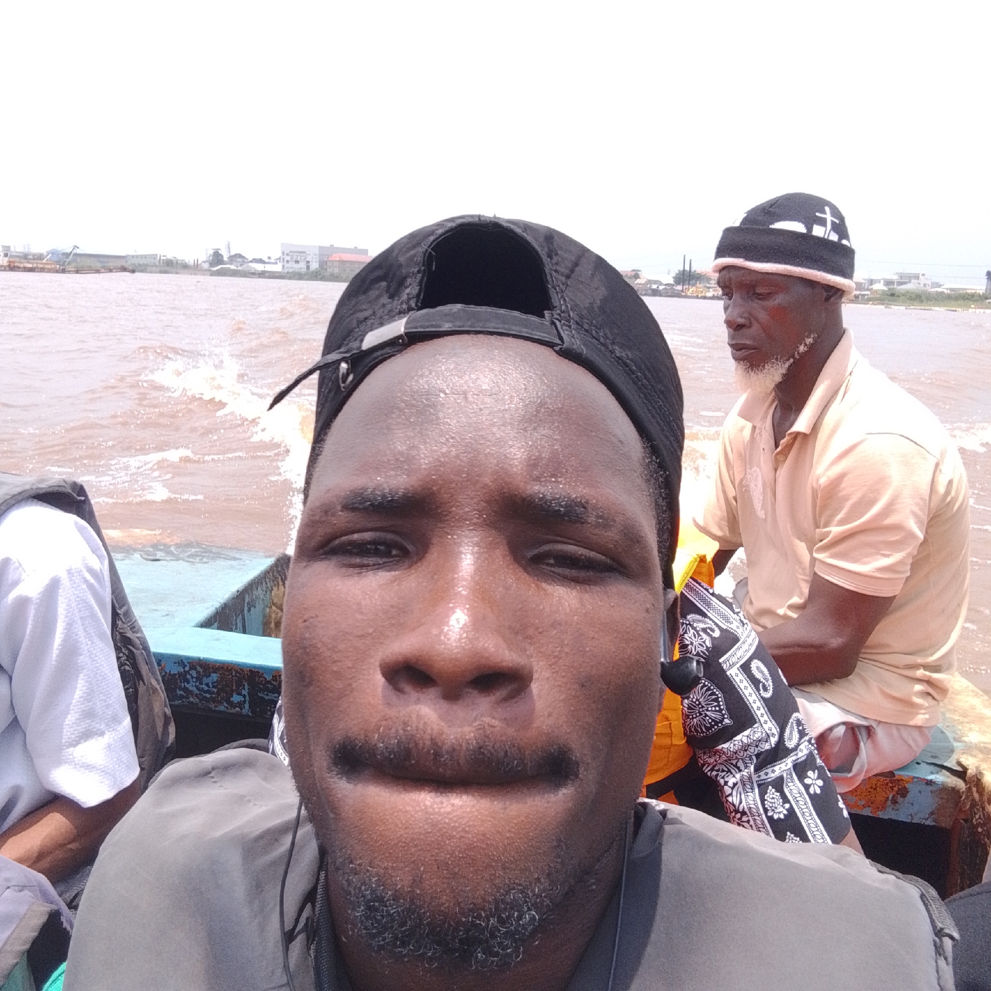 This is a selfie taken on a boat on the Lagos Lagoon in Ikorodu, Nigeria. In the foreground, a man wearing a black baseball cap and a grey shirt is looking at the camera. Behind him, another man wearing a tan polo shirt and a black and white beanie is seated. The boat is moving, creating ripples in the brown water. In the background, the Ikorodu skyline is visible, with industrial buildings and residential areas. The sky is overcast.