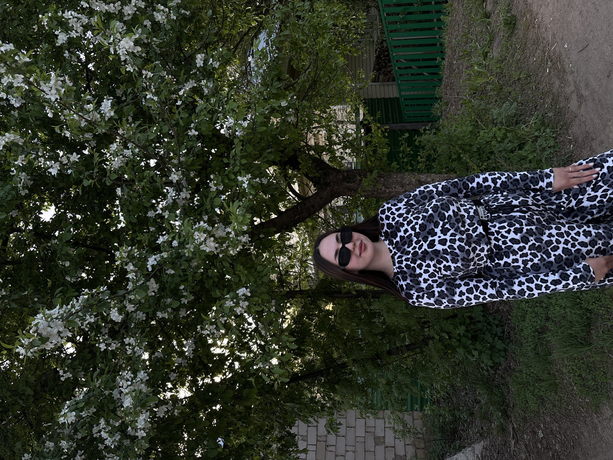 A woman wearing sunglasses and a black and white patterned outfit is standing outside near a tree with white blossoms. The location appears to be a residential area in Фоки, Russia, as evidenced by the presence of a green fence and a brick walkway. The woman is the primary subject of the image.