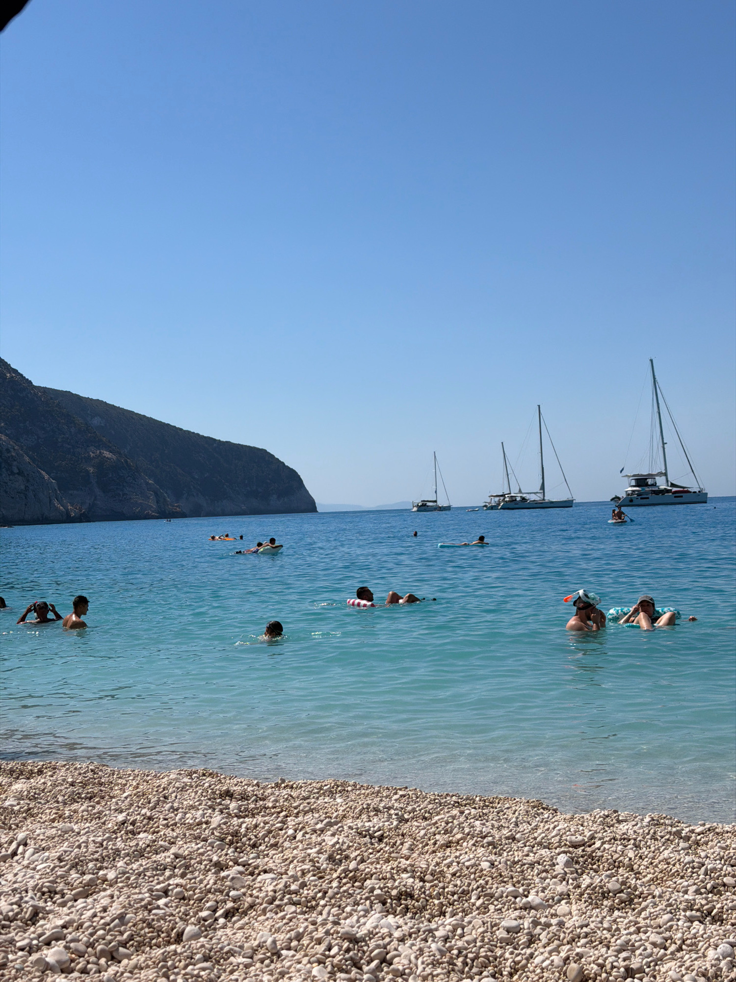 This image depicts a coastal scene at a beach, likely in the Municipal Unit of Apollonioi, Greece, characterized by its turquoise waters and a pebble shoreline. In the foreground, the beach is covered with a dense layer of light-colored pebbles. The sea is clear and blue, with several people enjoying various water activities. Some individuals are swimming, while others are floating on inflatable rings or rafts. In the mid-ground, a group of people are in the water, some appearing to be swimming, others relaxing on floats. Further out in the water, several sailboats are anchored, adding to the serene atmosphere. On the left side of the image, a steep, rocky cliff face rises from the sea, covered with sparse vegetation. The sky is a clear, bright blue, indicating a sunny day. The overall impression is one of a leisurely summer day at a Mediterranean beach.