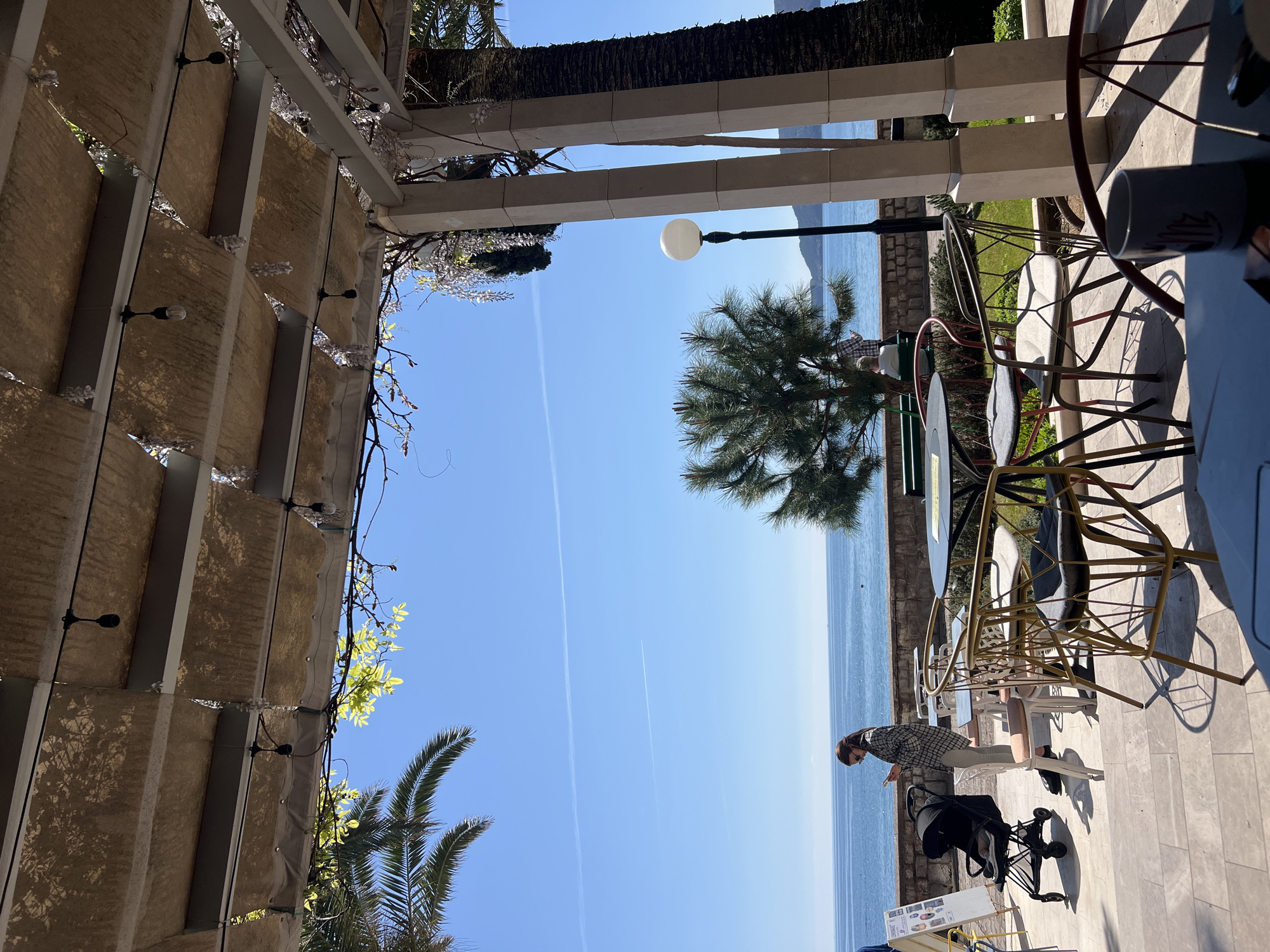 The image depicts an outdoor scene with a clear blue sky over the Adriatic Sea, likely in Rafailovići, Montenegro, given the context. The viewpoint appears to be from under a structure with a textured beige awning and a metal frame. To the right, there's outdoor seating with chairs and a table on a stone-tiled surface. A woman wearing a plaid shirt and sunglasses stands near a baby stroller next to the seating area. A solitary street lamp and a tree break the horizon line. The sky is clear, with two visible contrails from aircraft.