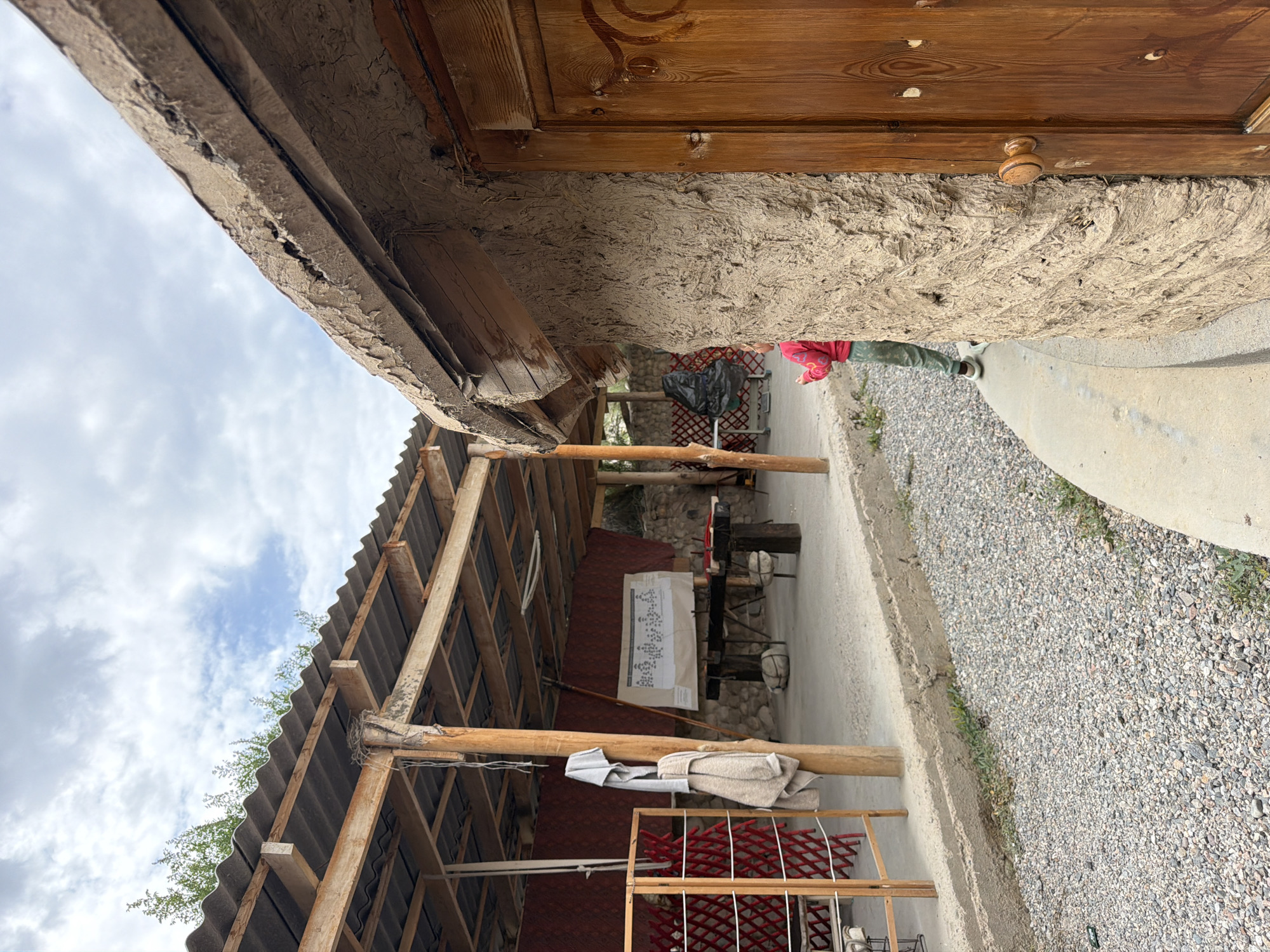 A medium shot captures a scene in Кызыл-Туу, Kyrgyzstan. In the foreground, the textured, mud-plastered wall of a rustic building with a wooden door is visible on the right. A child in a pink shirt and green pants stands with their back to the camera, looking towards a covered outdoor area. This area, likely a workshop or gathering space, features a corrugated metal roof supported by wooden beams and poles. Hanging from a pole are two pieces of clothing. A whiteboard with diagrams is displayed on the back wall of the covered area, and a rough wooden table stands in front of it. The ground is covered with gravel and some sparse green vegetation. In the background, a wooden lattice structure and a black plastic bag are present, with a stone wall and trees visible further back. The sky above is cloudy.