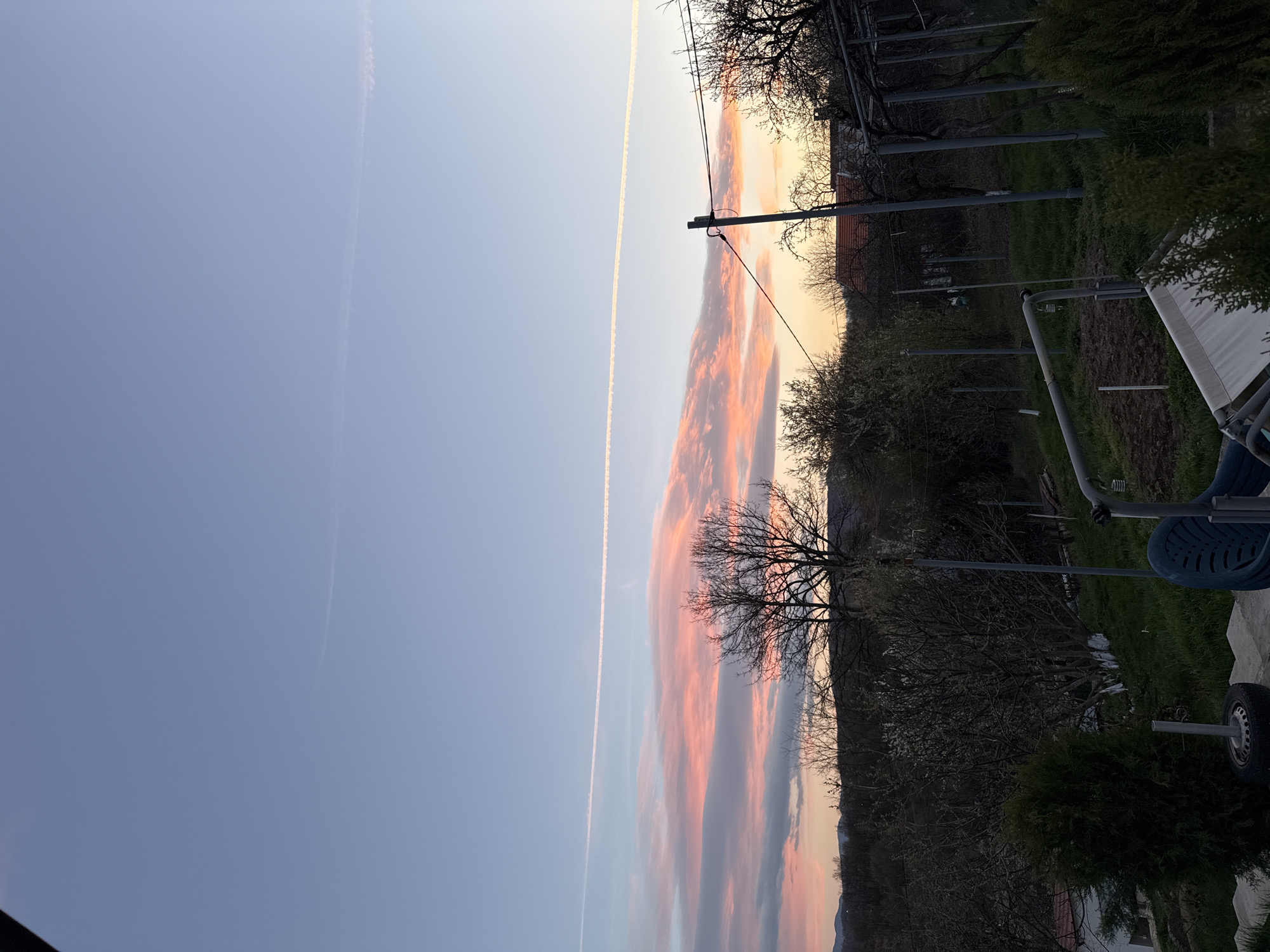 The image shows a landscape with a prominent sky. On the left side of the frame is a blue sky with a contrail. The right side of the frame displays a colorful sky with orange and gray clouds. The lower portion of the frame shows a rural setting, including trees, poles, a chair, and a tire. The overall impression is of a tranquil outdoor scene with a vivid sky. This location in the city of Balinovtsi, Bulgaria.